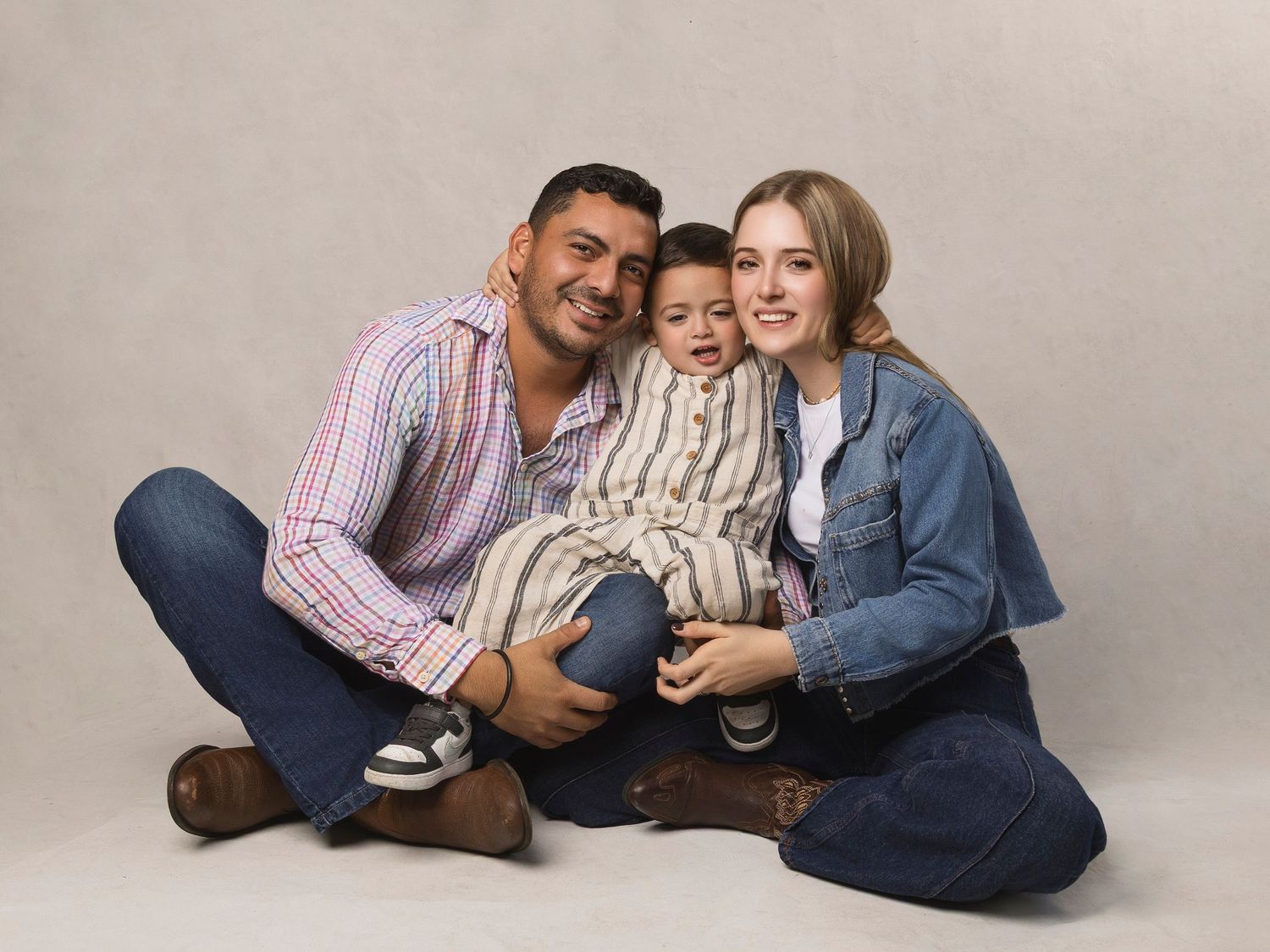 Smiling family of three sitting on floor together in casual studio portrait session.