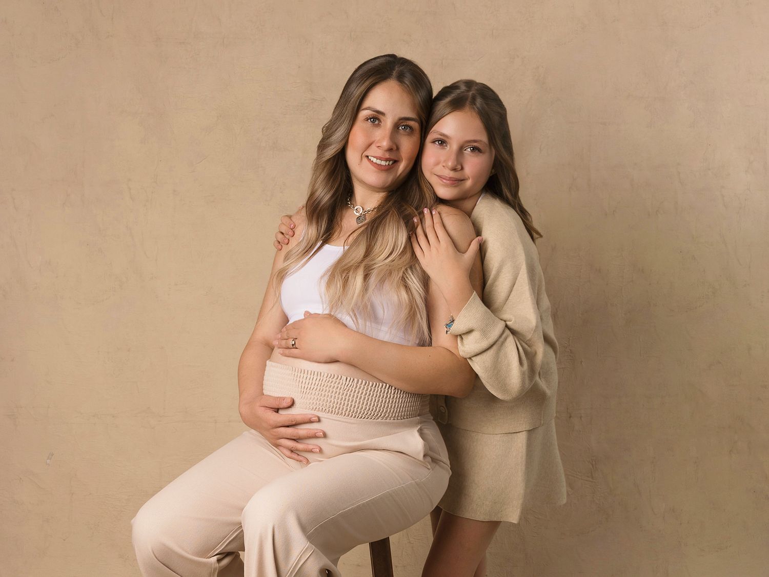 Pregnant woman sitting on a stool with a teenage girl leaning against her in a studio portrait.
