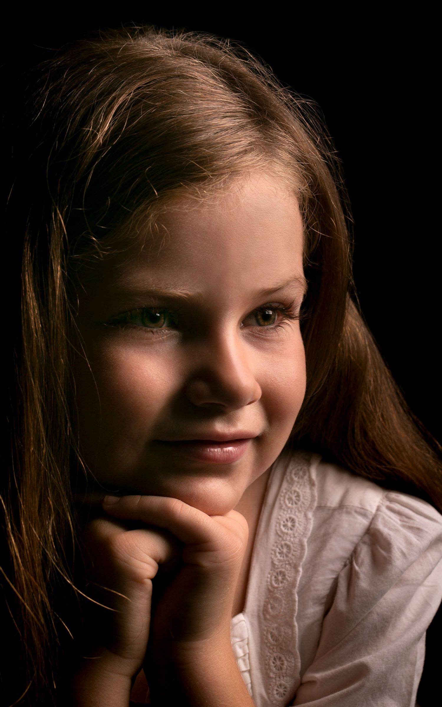 Young girl with long brown hair resting chin on hands, smiling softly in studio portrait.