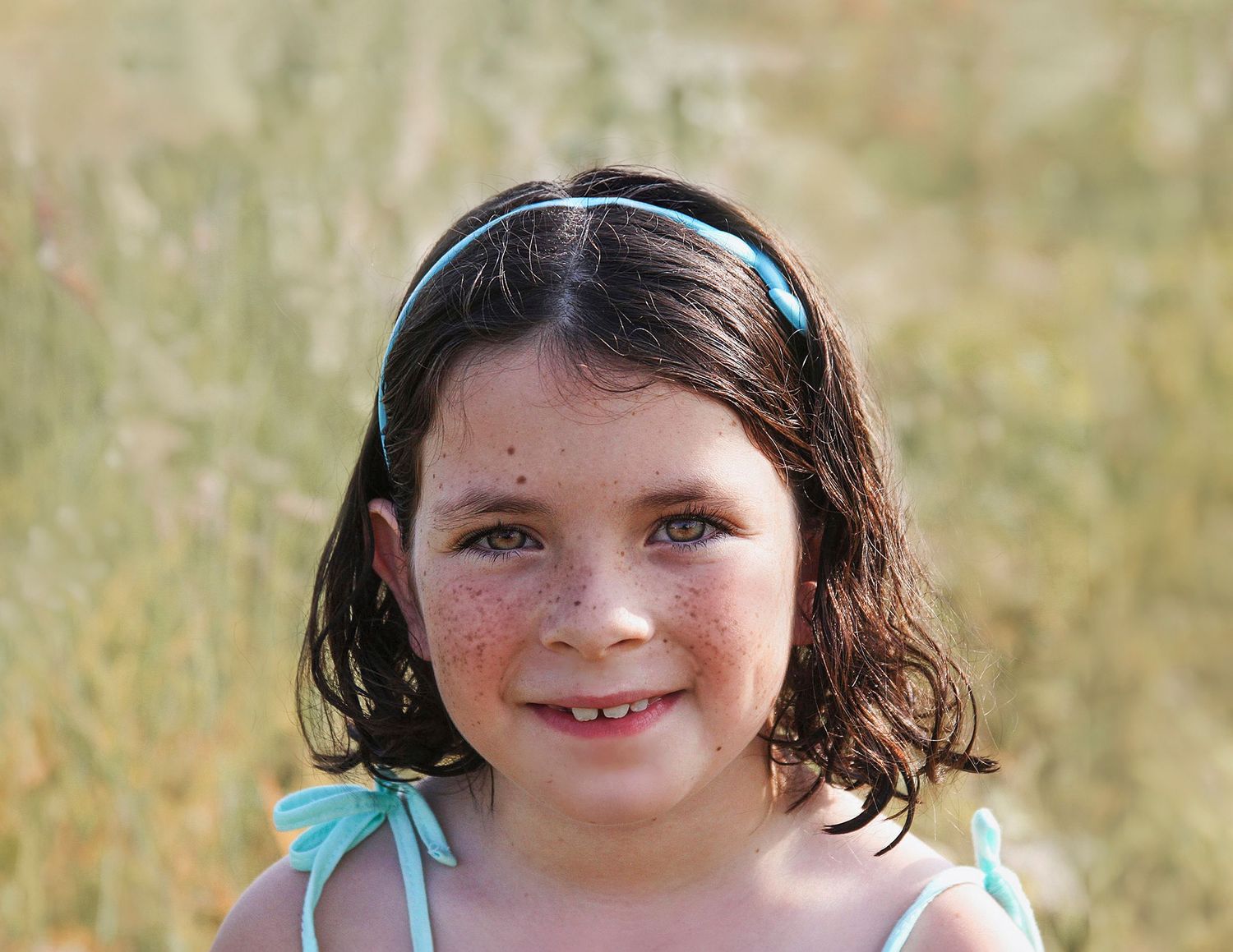 Young smiling girl with freckles and dark hair wearing a blue headband outdoors in a field.
