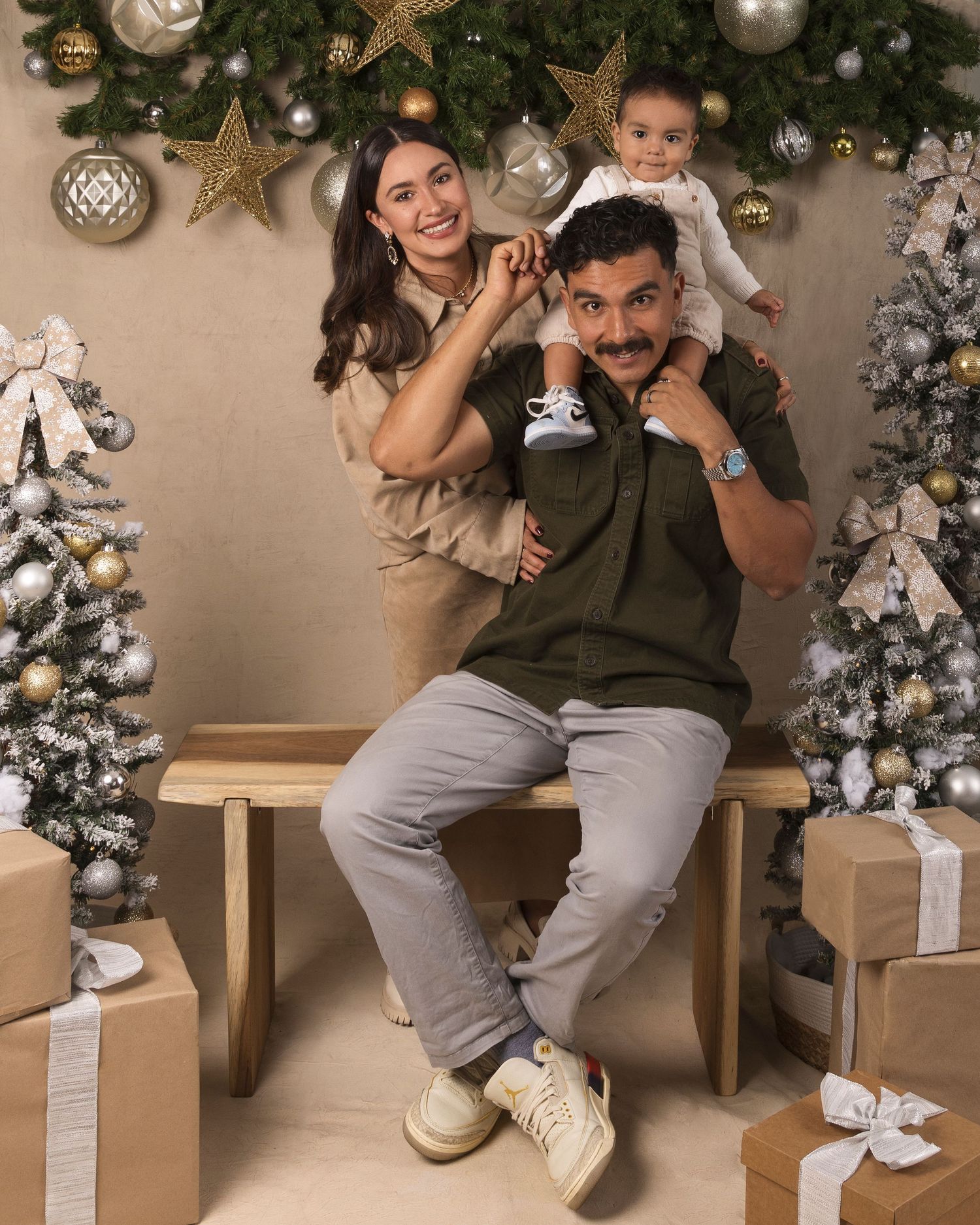 Family Christmas photo with parents and baby on wooden bench surrounded by decorated trees and gold ornaments.