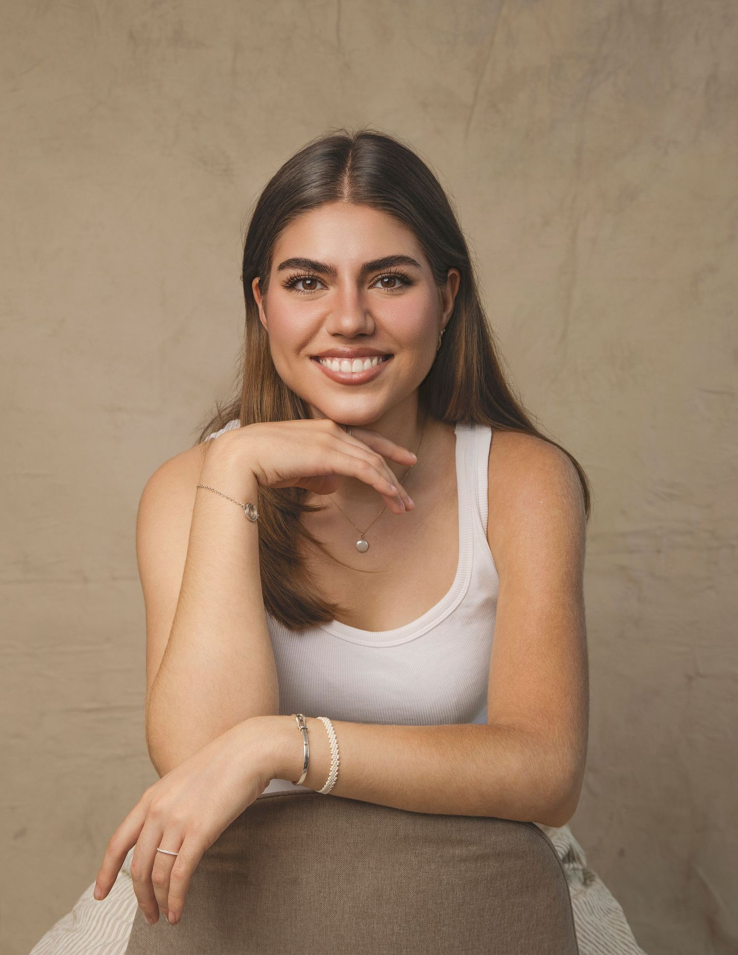 Young woman in white tank top smiling warmly while leaning forward against neutral studio backdrop.