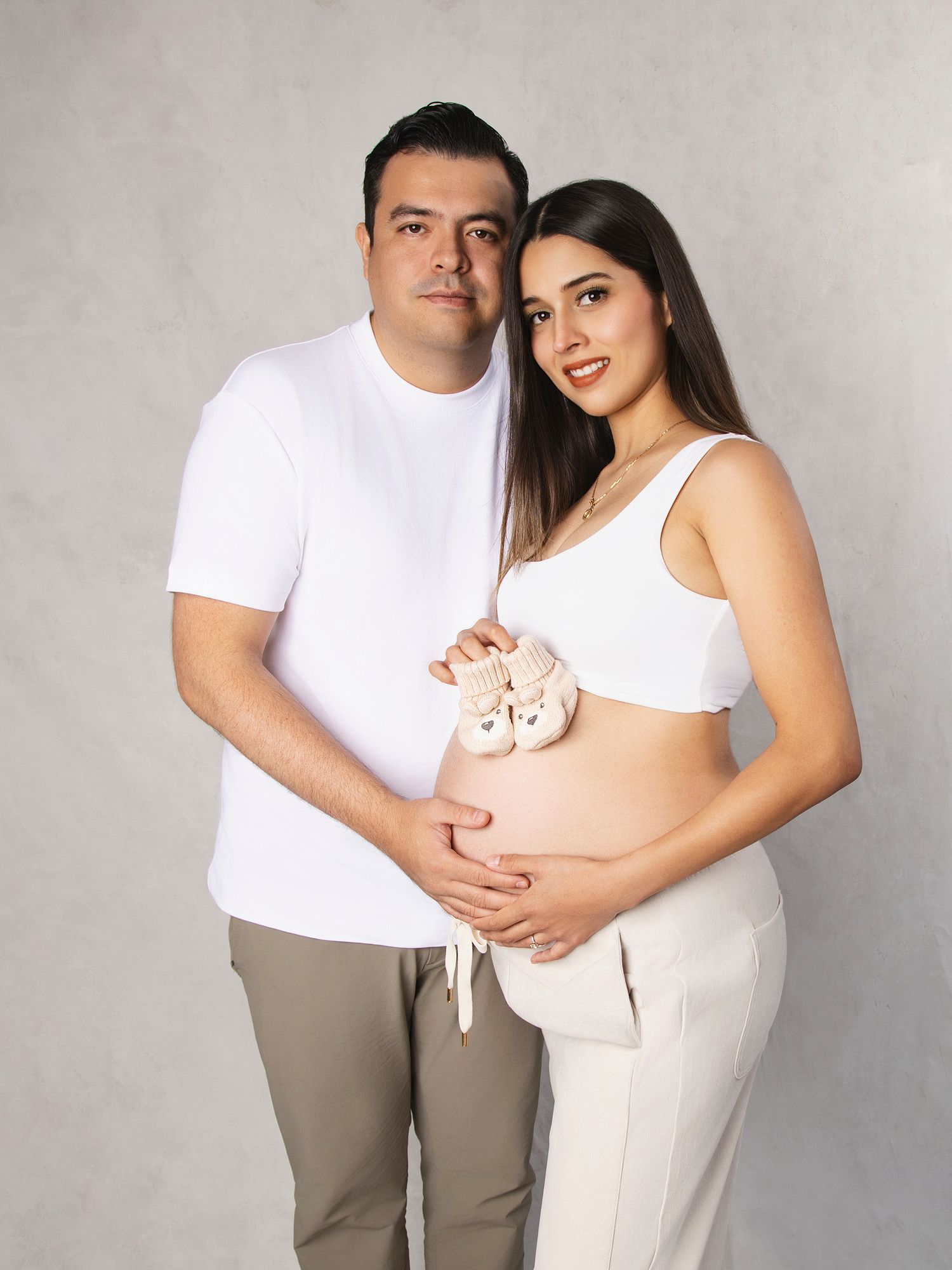Couple in white outfits holding baby shoes on pregnant belly in studio.