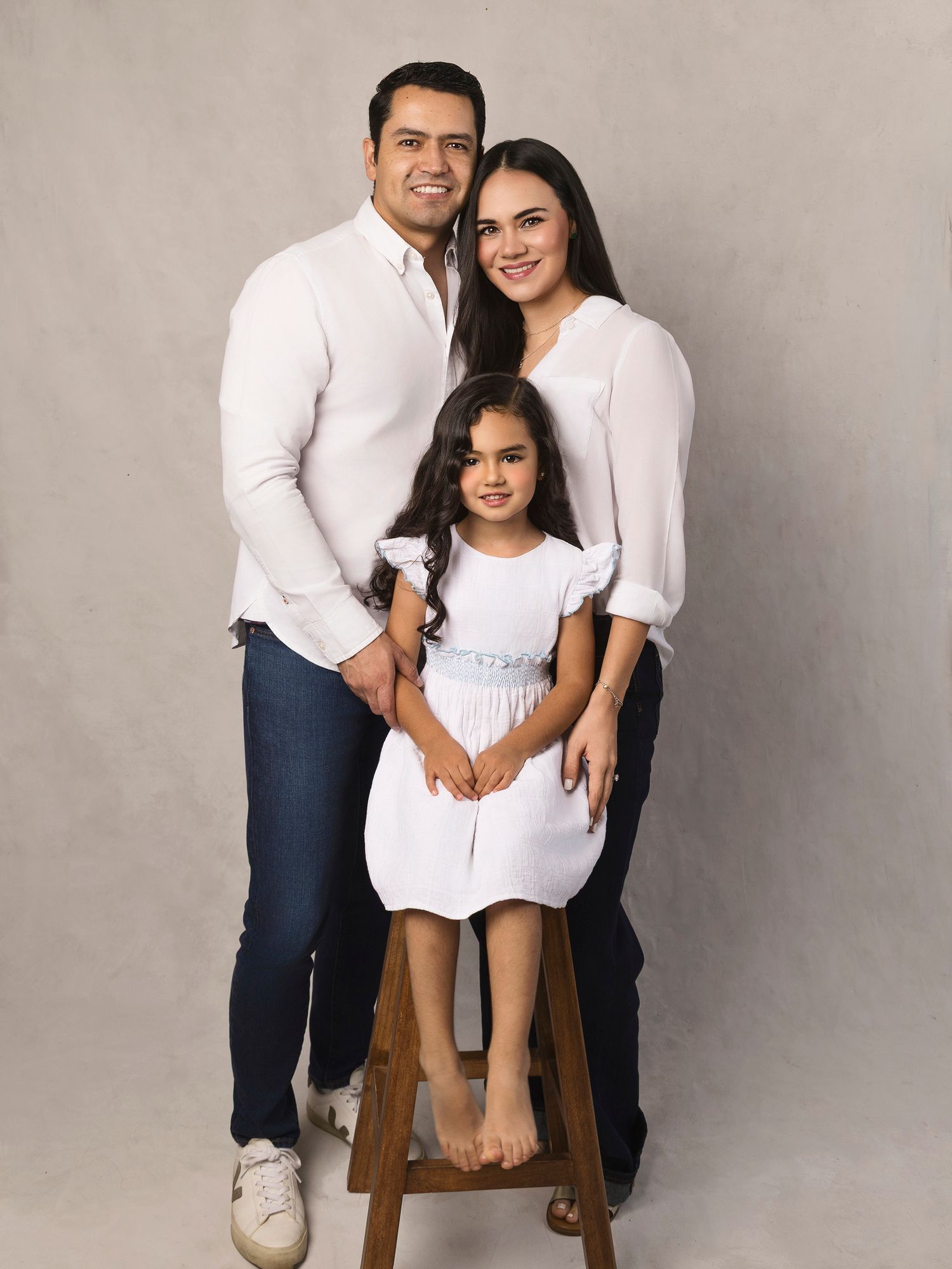Happy family of three in matching white outfits posing with young daughter on stool against gray studio backdrop.