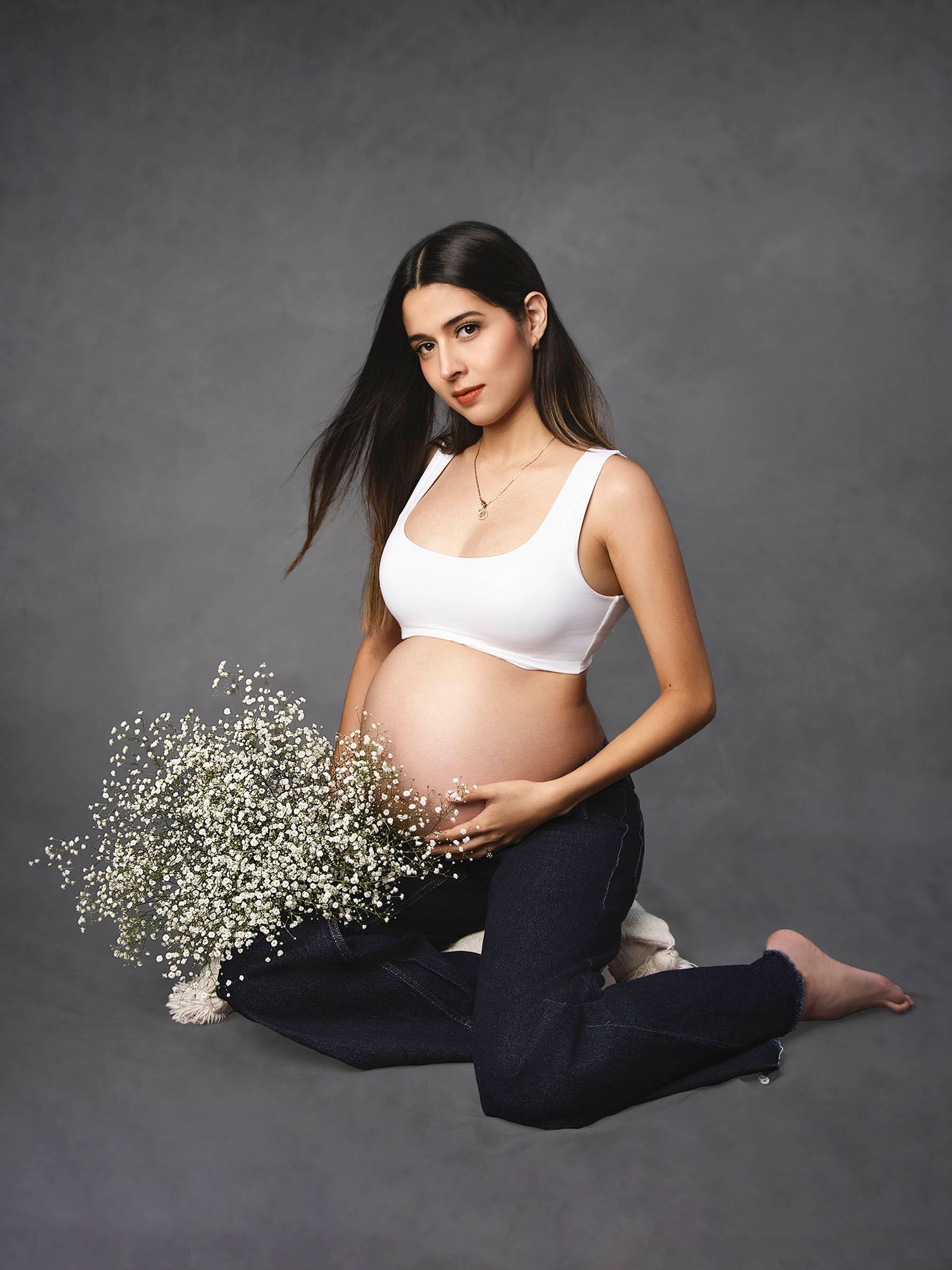 Pregnant woman in white crop top and dark jeans sits cross-legged holding baby bump with white wildflowers.