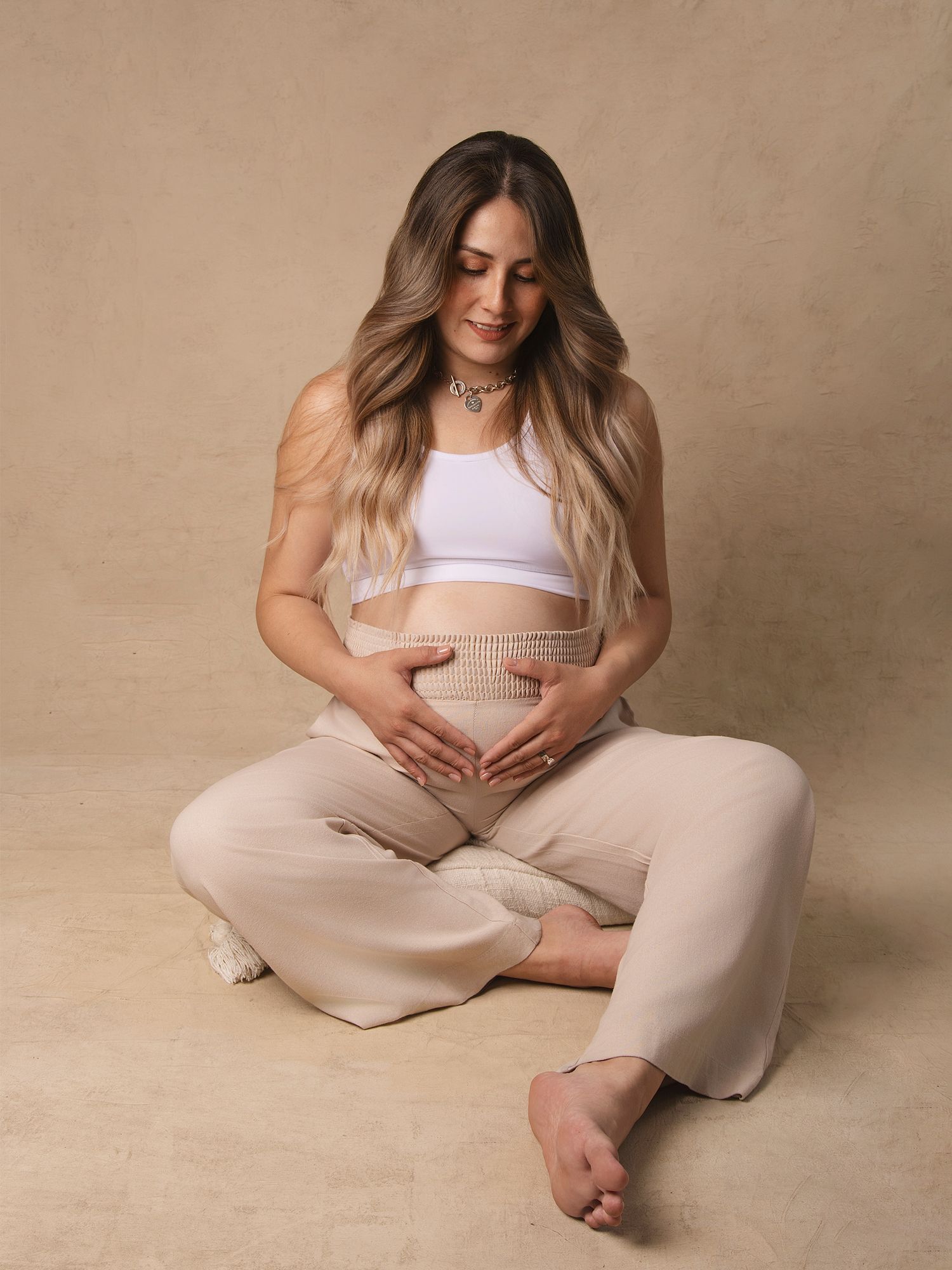 Pregnant woman in white crop top and beige pants sitting cross-legged cradling her baby bump in a studio.