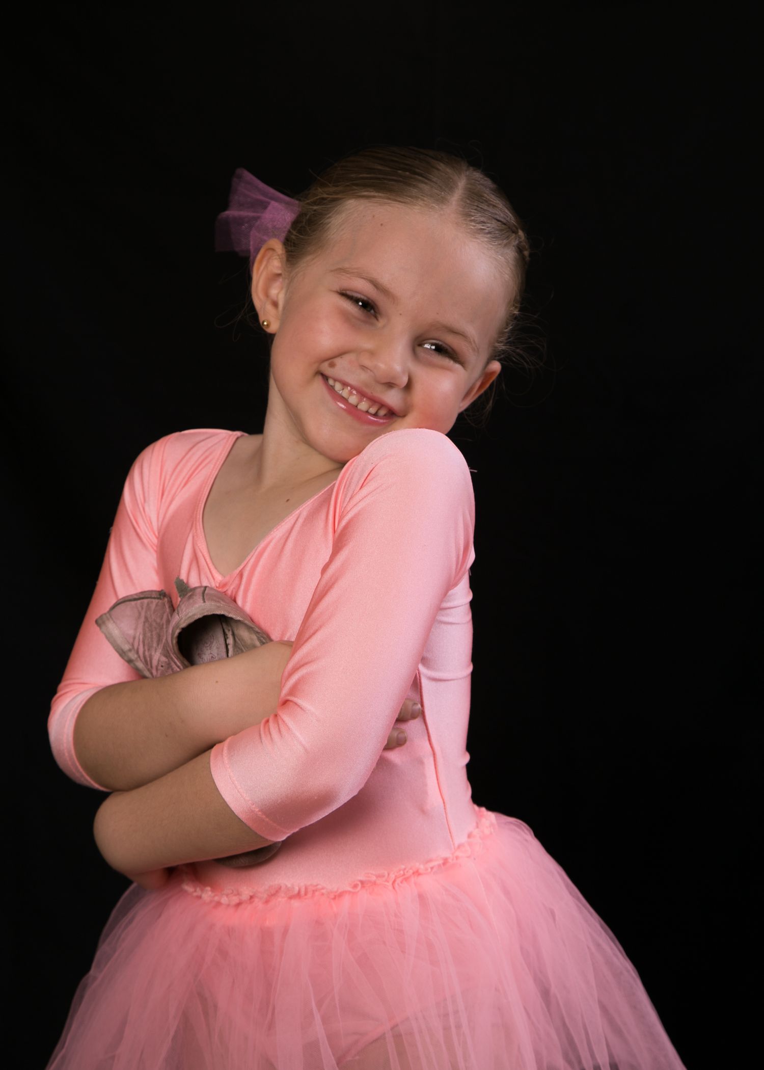 Young girl in a pink ballet tutu smiling with arms crossed against a black background.