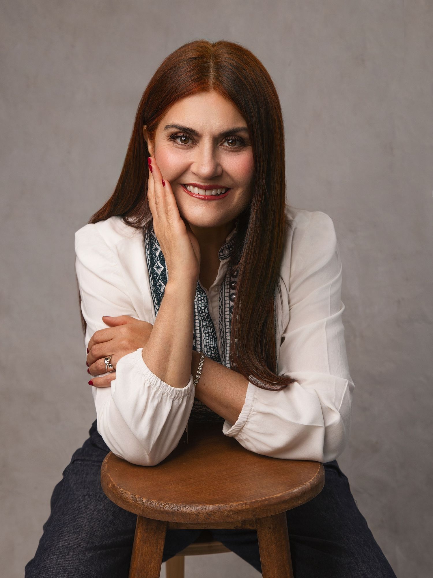 Smiling woman with long brown hair in white blouse sits on wooden stool, posing with hand on cheek.