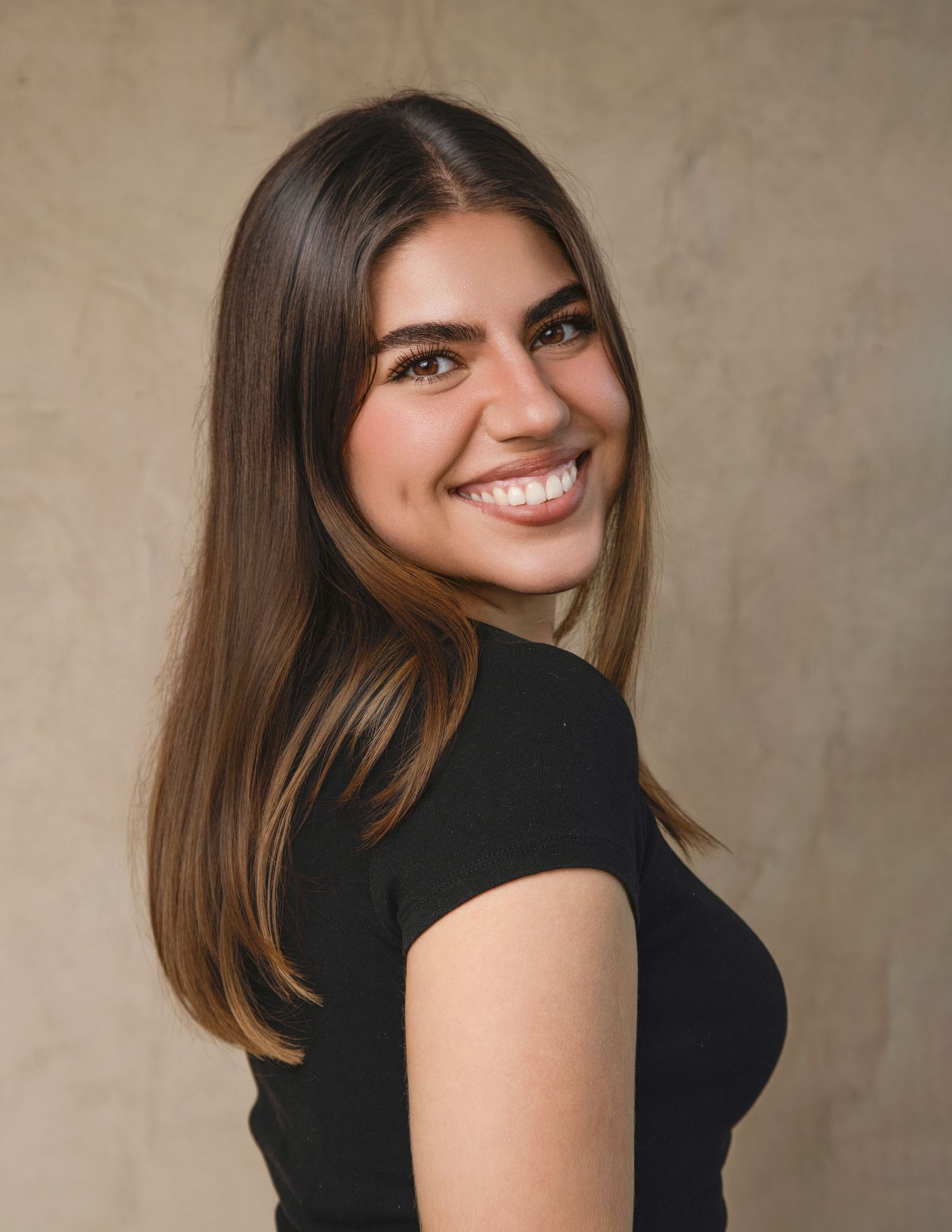 Side profile portrait of smiling young woman with brown highlighted hair against warm beige backdrop.