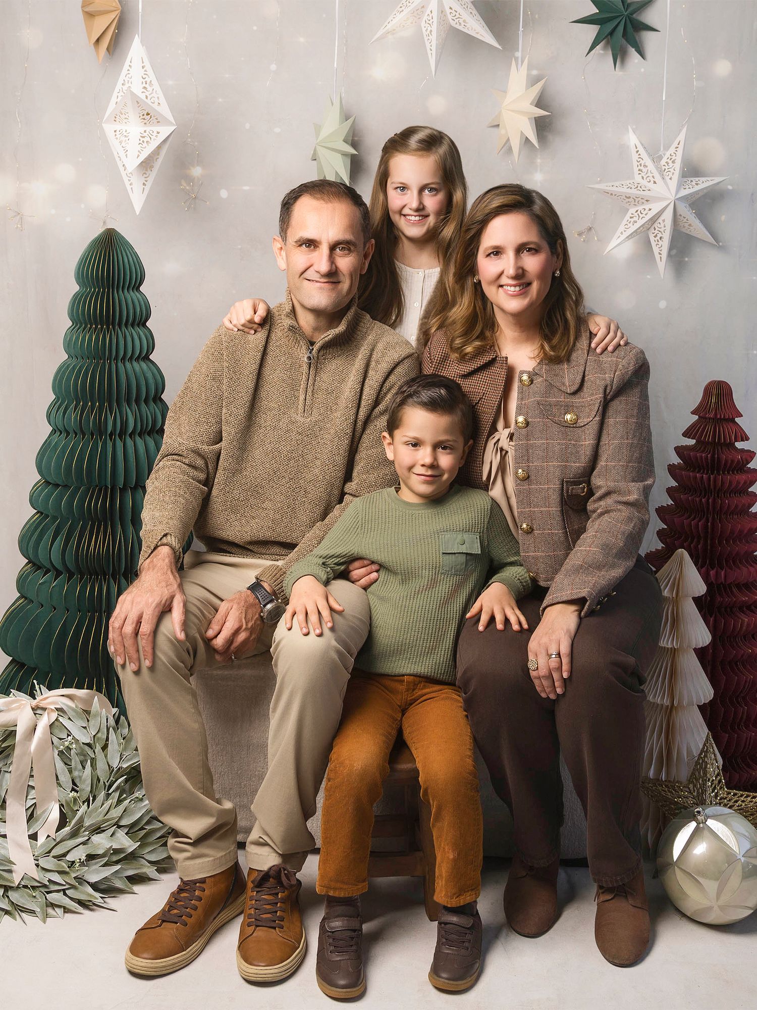 Festive family Christmas photo with parents and two children posing among holiday trees and decor.