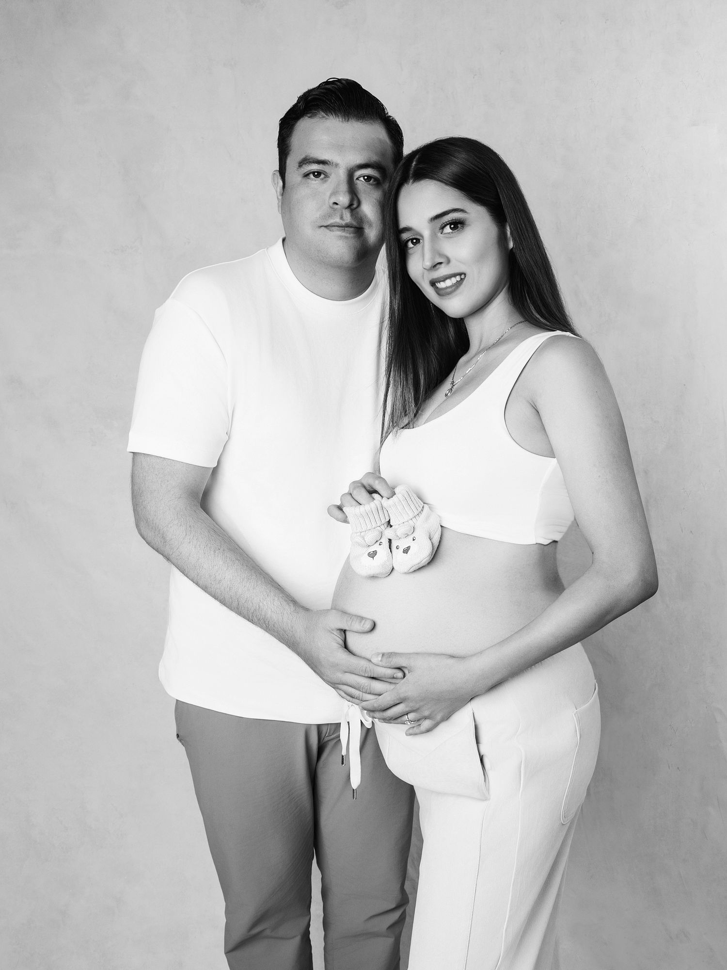 Black and white maternity photo of a couple holding baby shoes over a pregnant belly in a studio.
