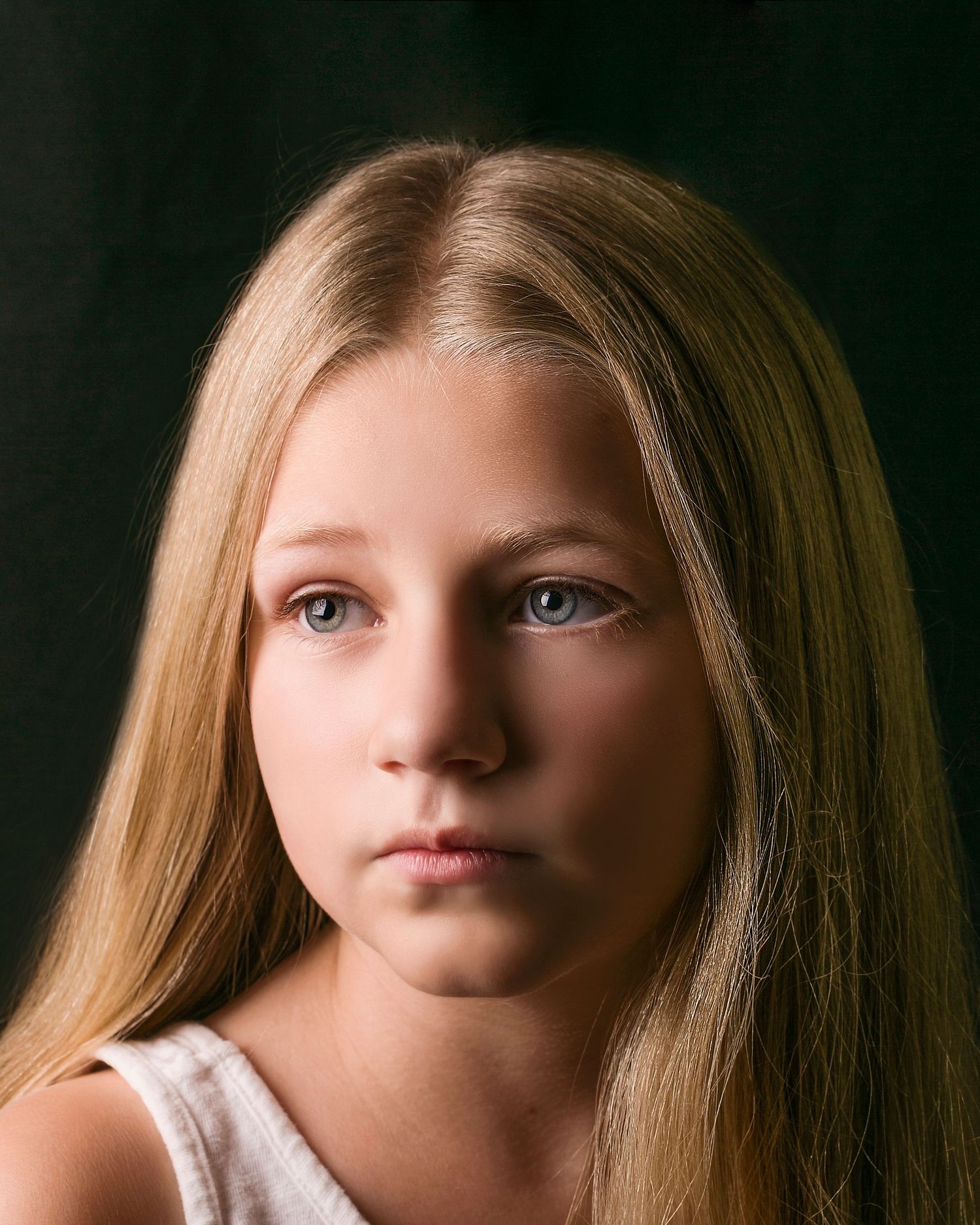 Young blonde girl with blue eyes in serious close-up portrait against dark dramatic background.