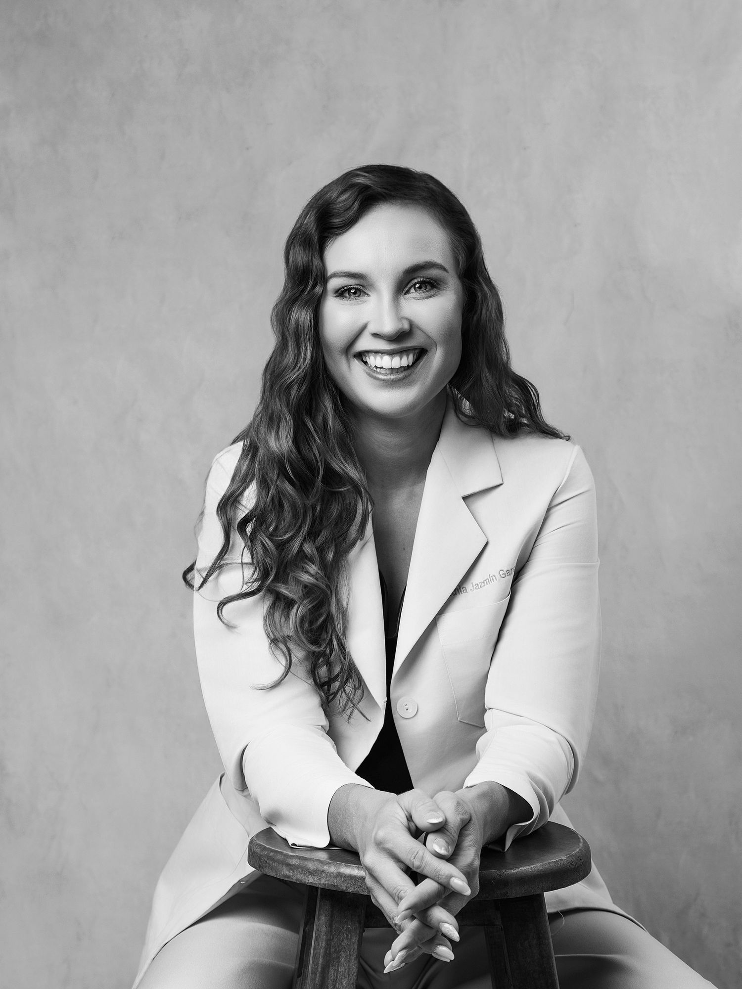 Black and white professional headshot of a smiling woman in a blazer, seated with hands clasped.