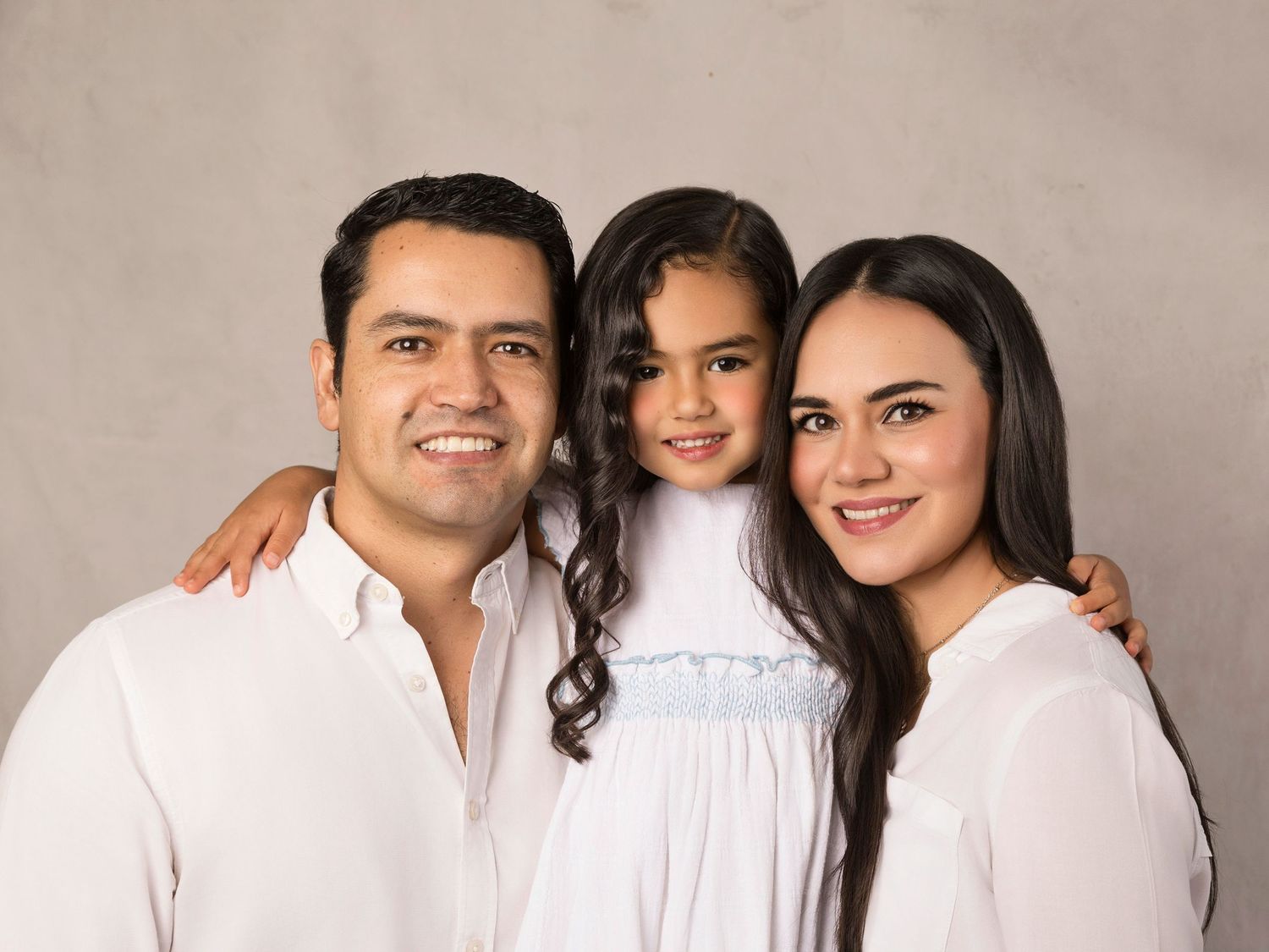 Happy family of three in white outfits posing together in a bright studio portrait.