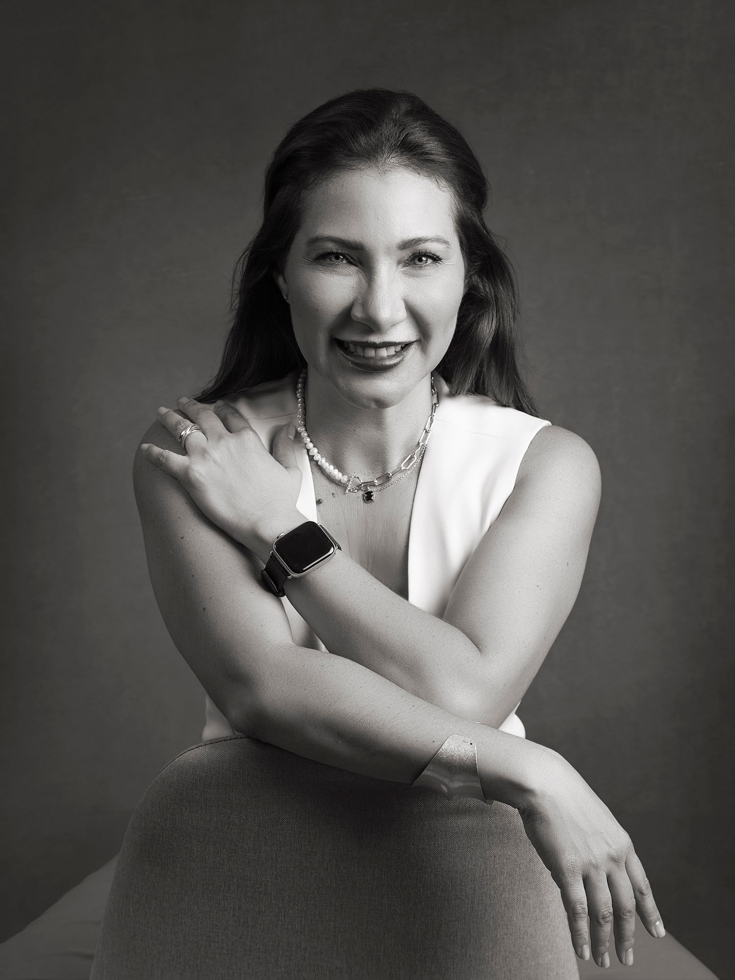 Black and white studio portrait of a smiling woman in a white dress wearing a smartwatch and pearl necklace.