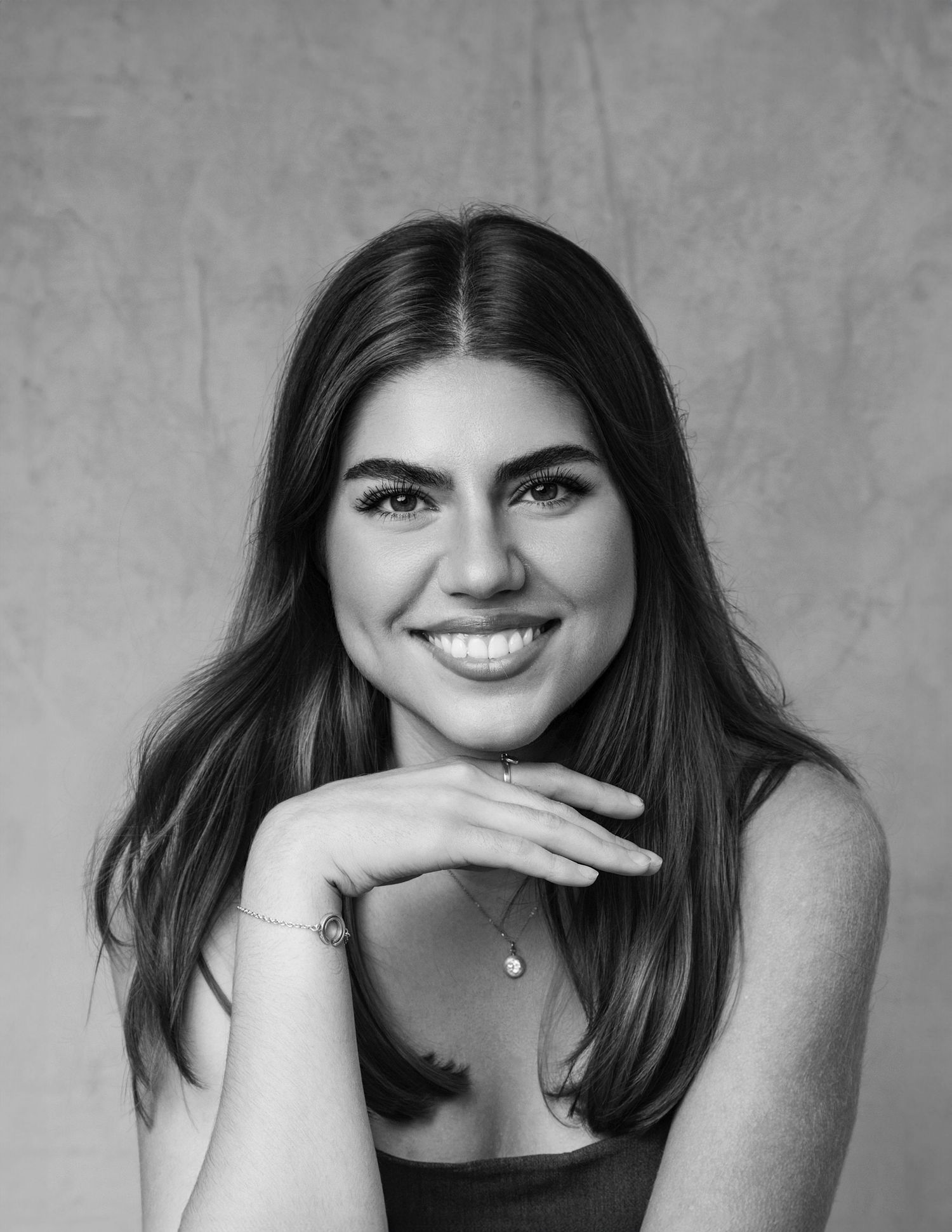 Black and white close-up portrait of smiling young brunette woman with hands folded under chin.