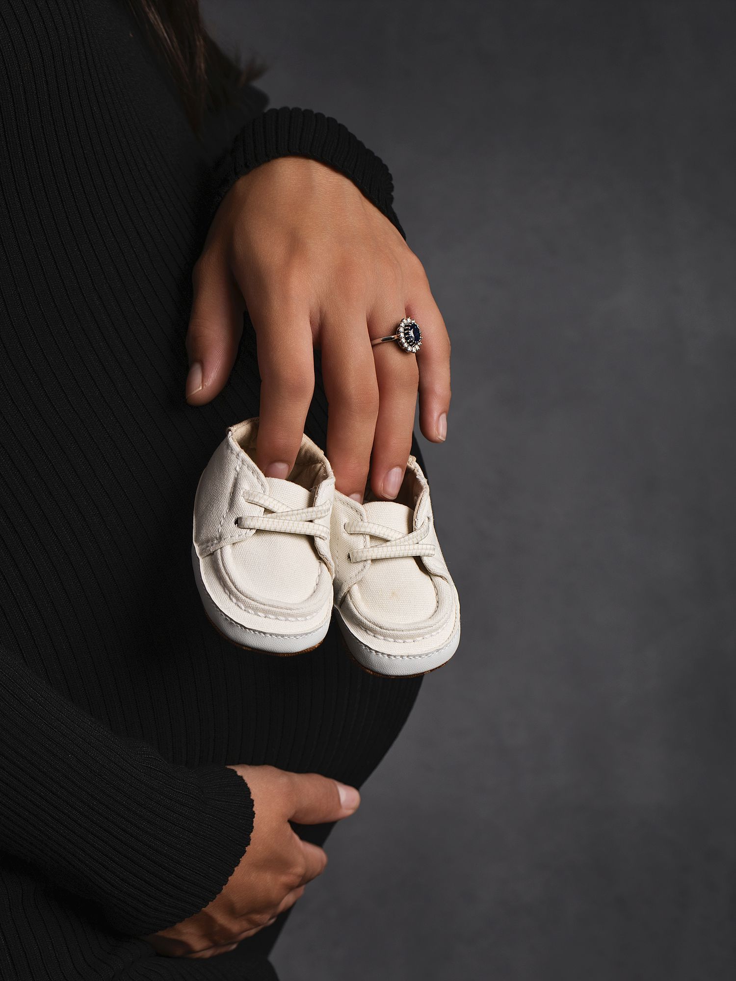 Close-up of pregnant woman in black dress holding tiny white baby loafers over her baby bump.
