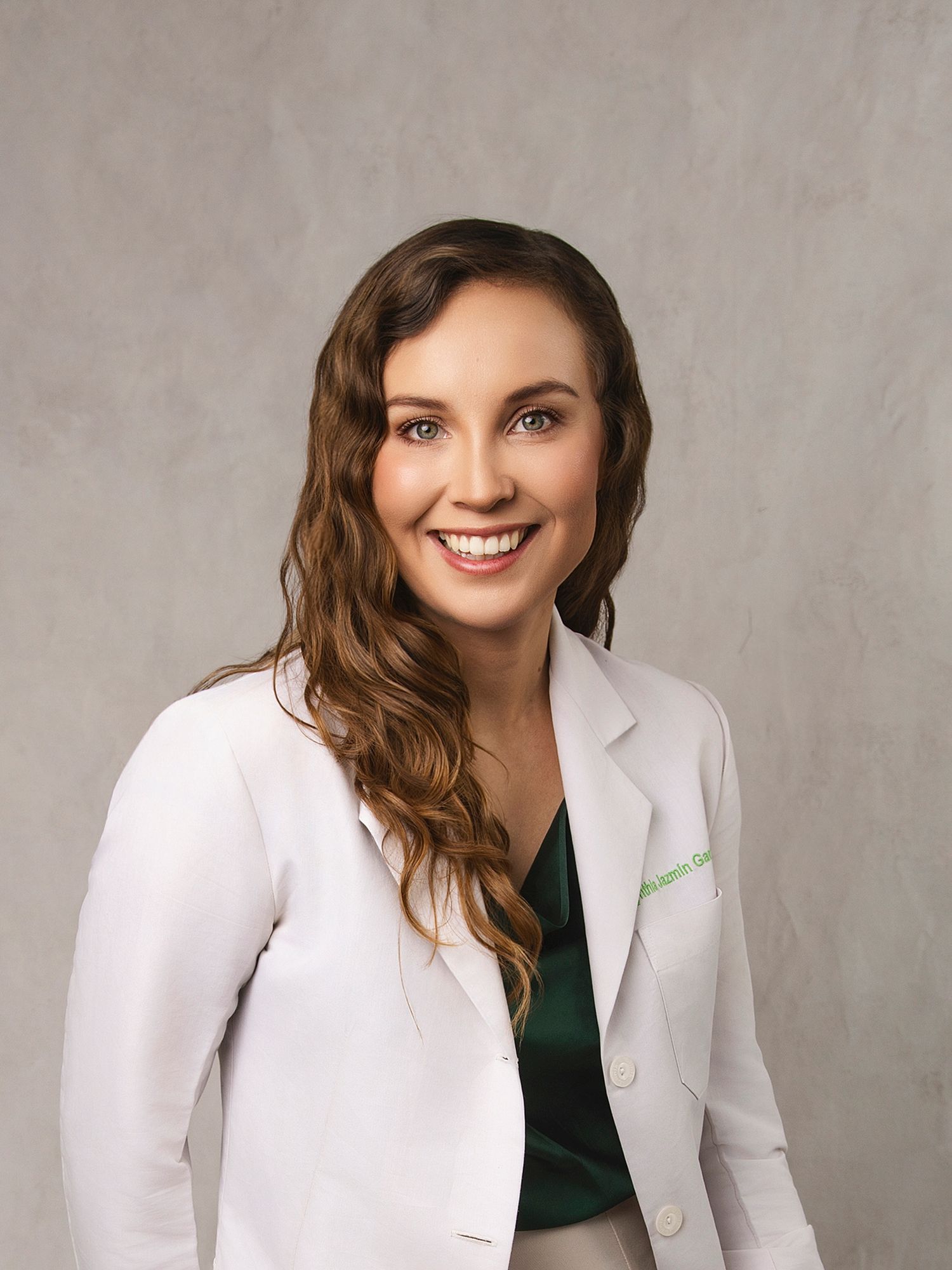 Smiling woman in white lab coat and green top, professional healthcare portrait against gray background.