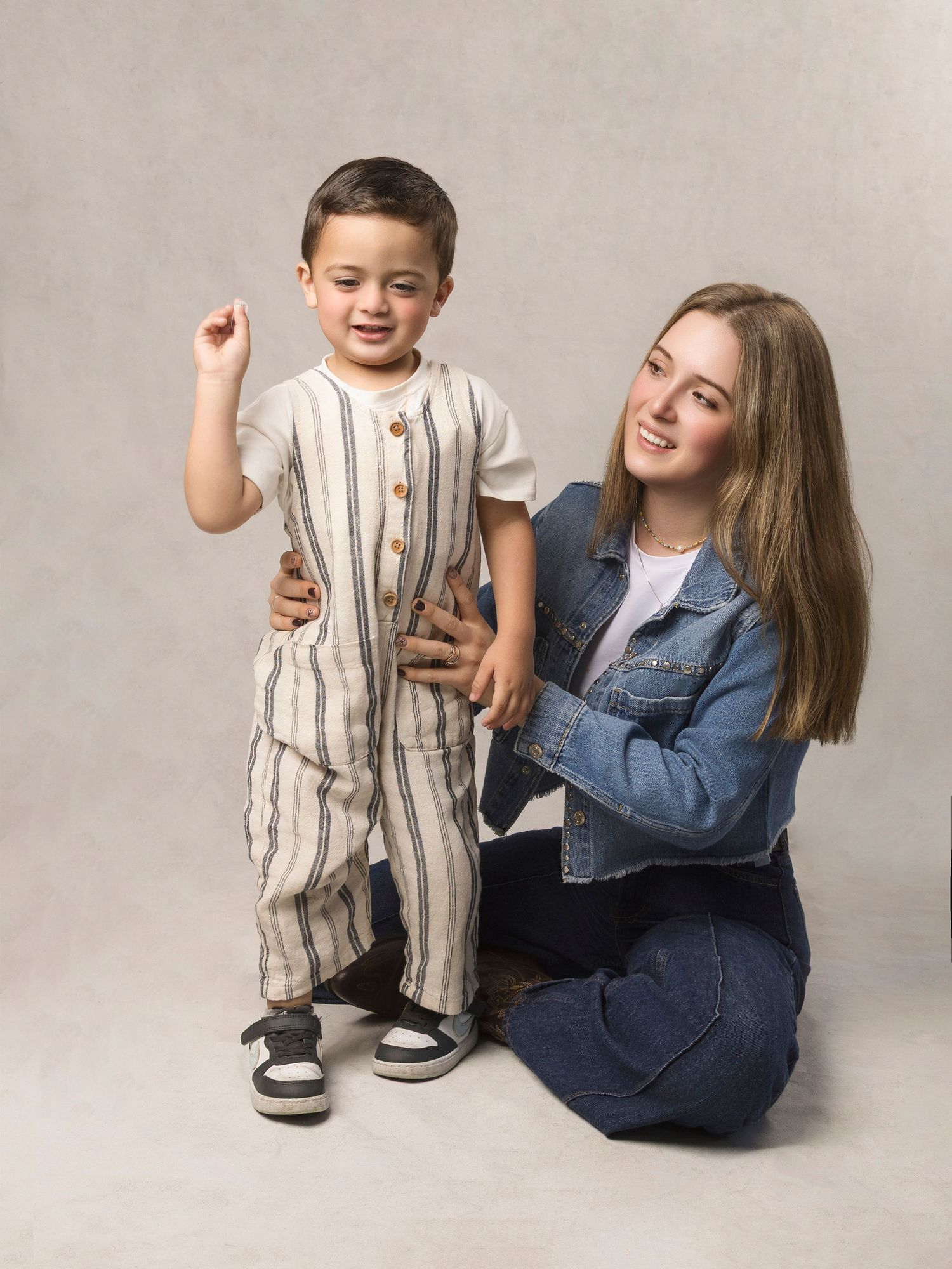 Young woman kneeling beside cheerful toddler boy in striped outfit in studio portrait.