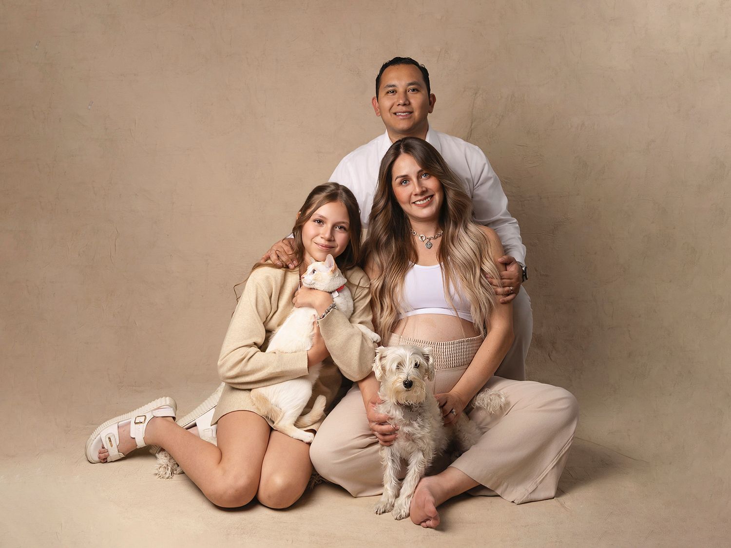 Family maternity photo with pregnant mother, father, teenage daughter, and a small white dog in a studio.