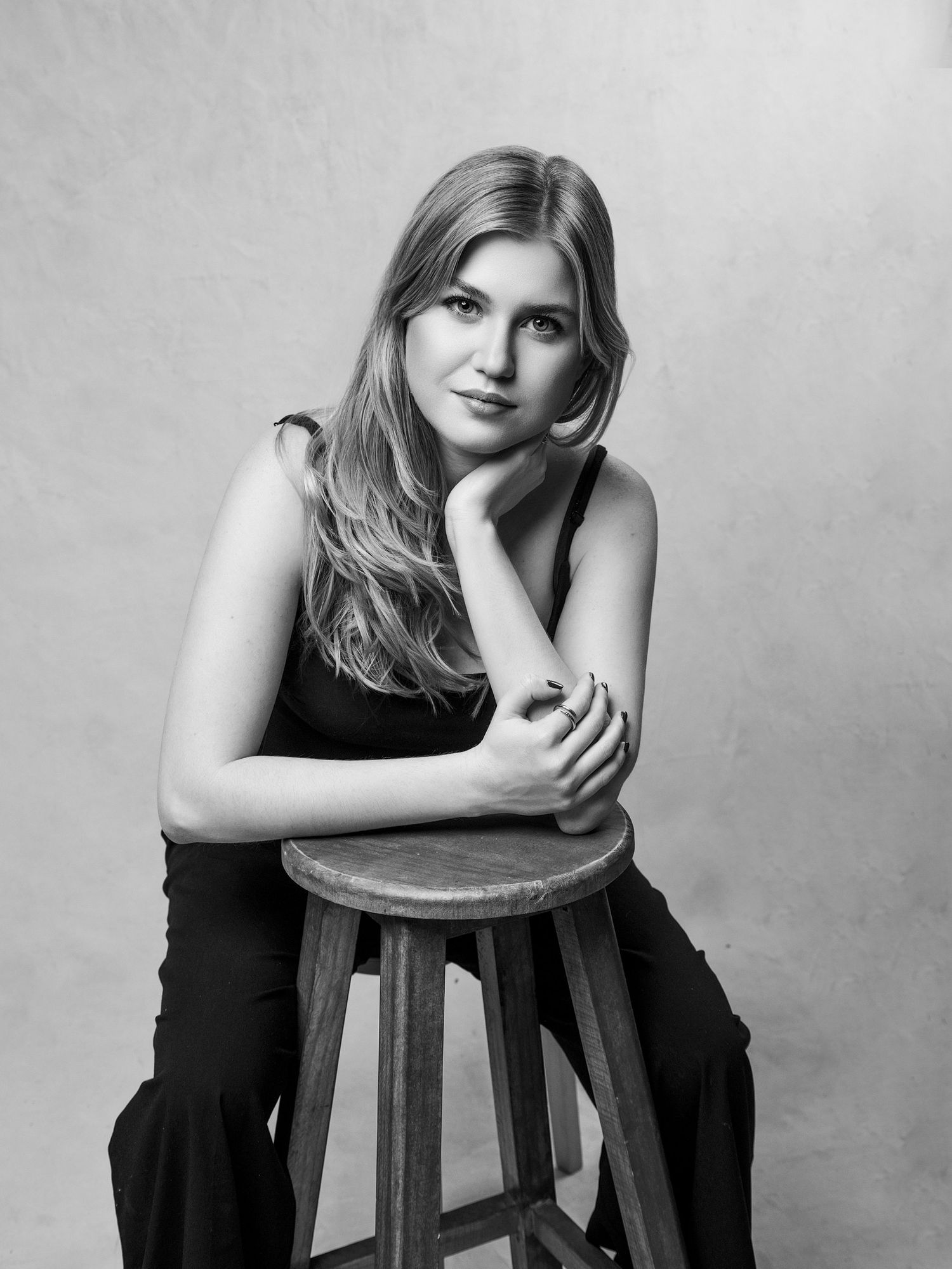 Black and white studio portrait of a young woman in black outfit leaning on a wooden stool.