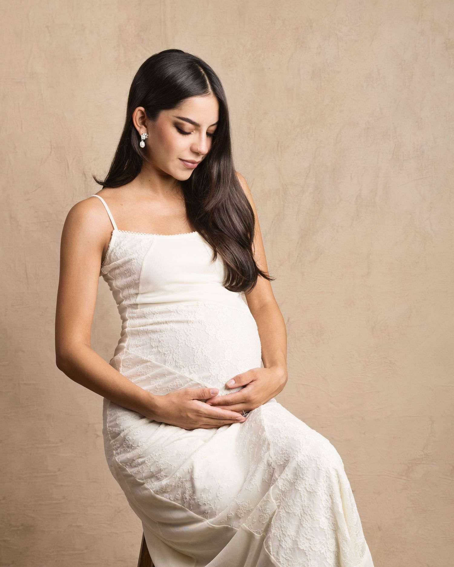 Elegant pregnant woman in white lace gown sits gracefully holding her baby bump in a studio portrait.