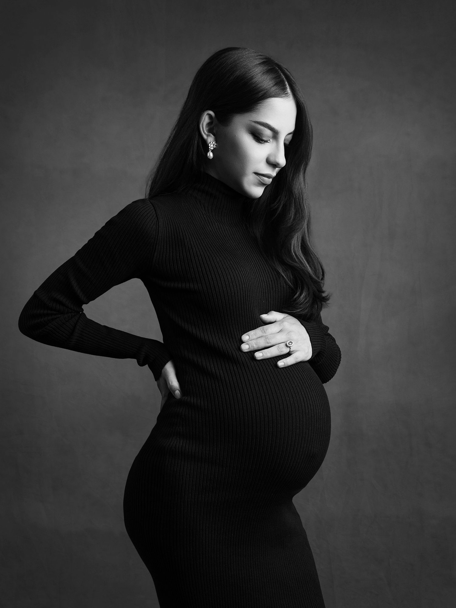 Elegant black and white maternity portrait of a woman in a black turtleneck dress cradling her baby bump.