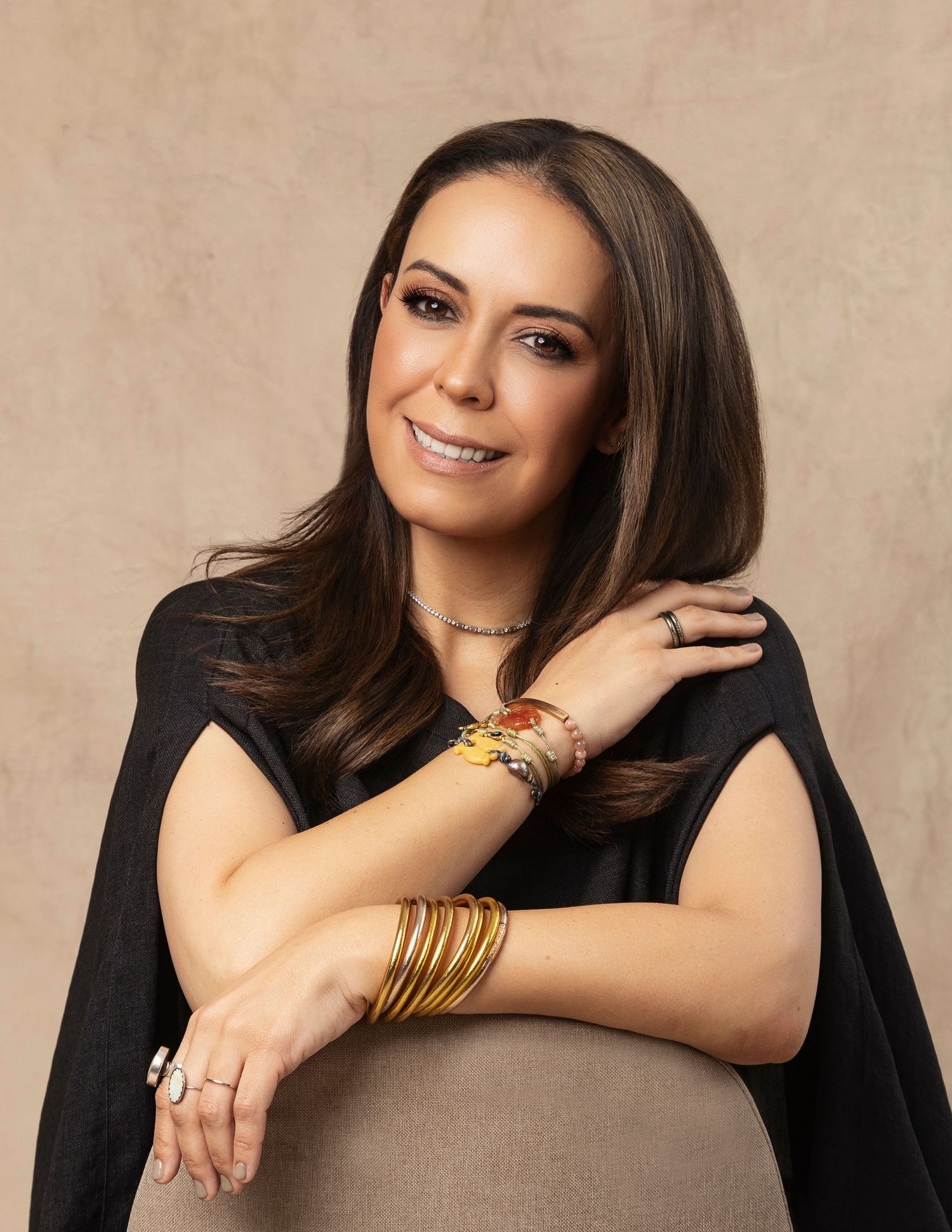 Woman in black cape top wearing gold bangles and jewelry posing elegantly against beige background.