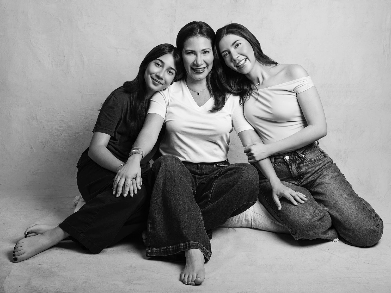 Black and white studio portrait of three women sitting together, smiling warmly in casual outfits.