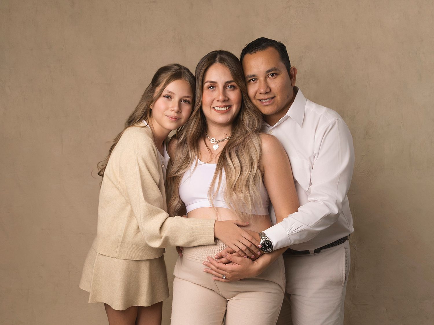 Family maternity portrait with pregnant mother, father, and daughter embracing in a warm studio setting.