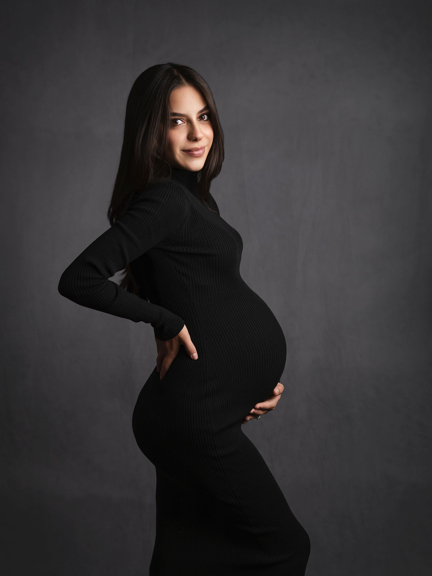Pregnant woman in fitted black turtleneck dress poses sideways showing her baby bump in a studio portrait.