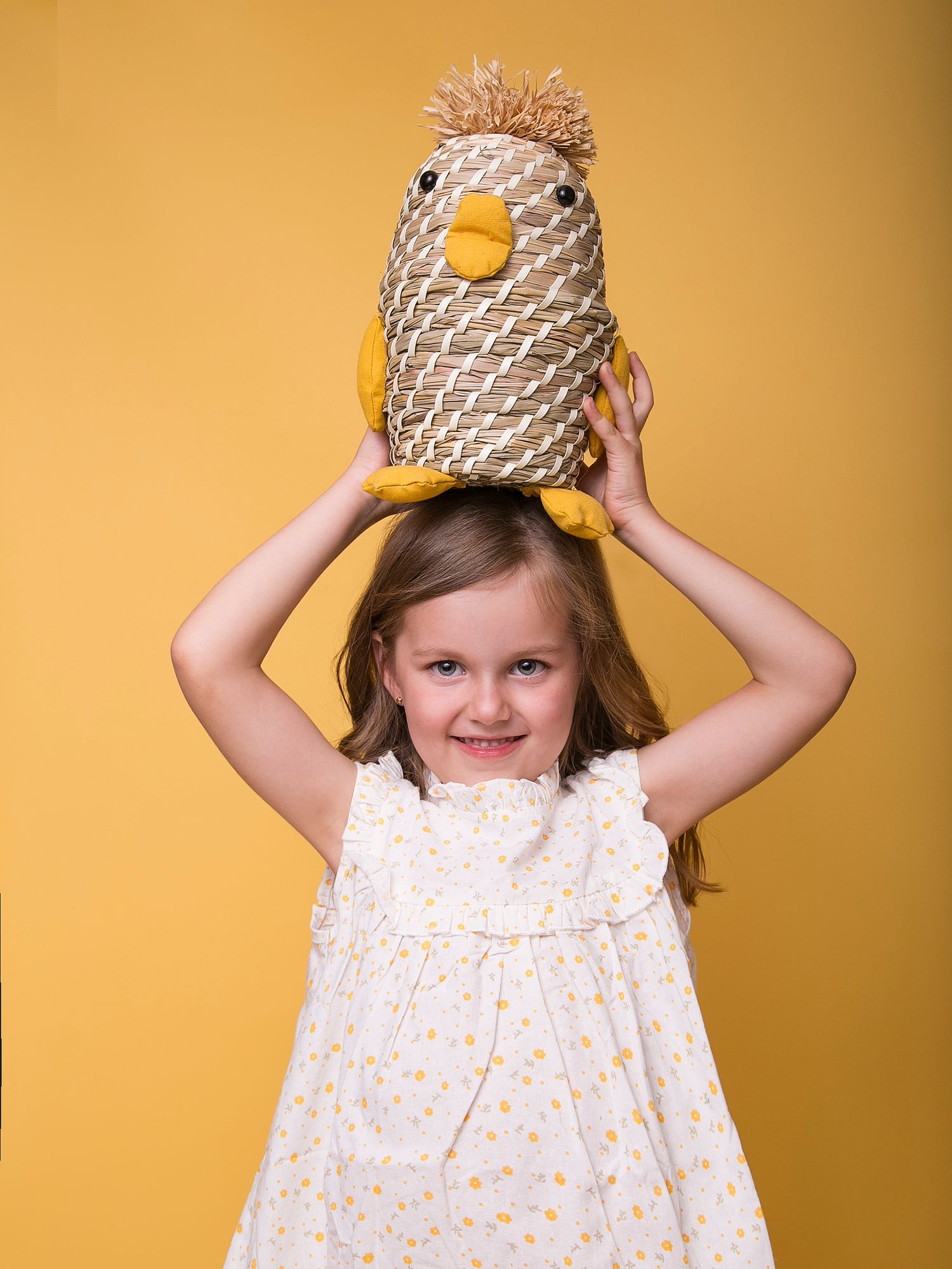Smiling young girl balancing woven chick toy on head wearing daisy dress on yellow background.