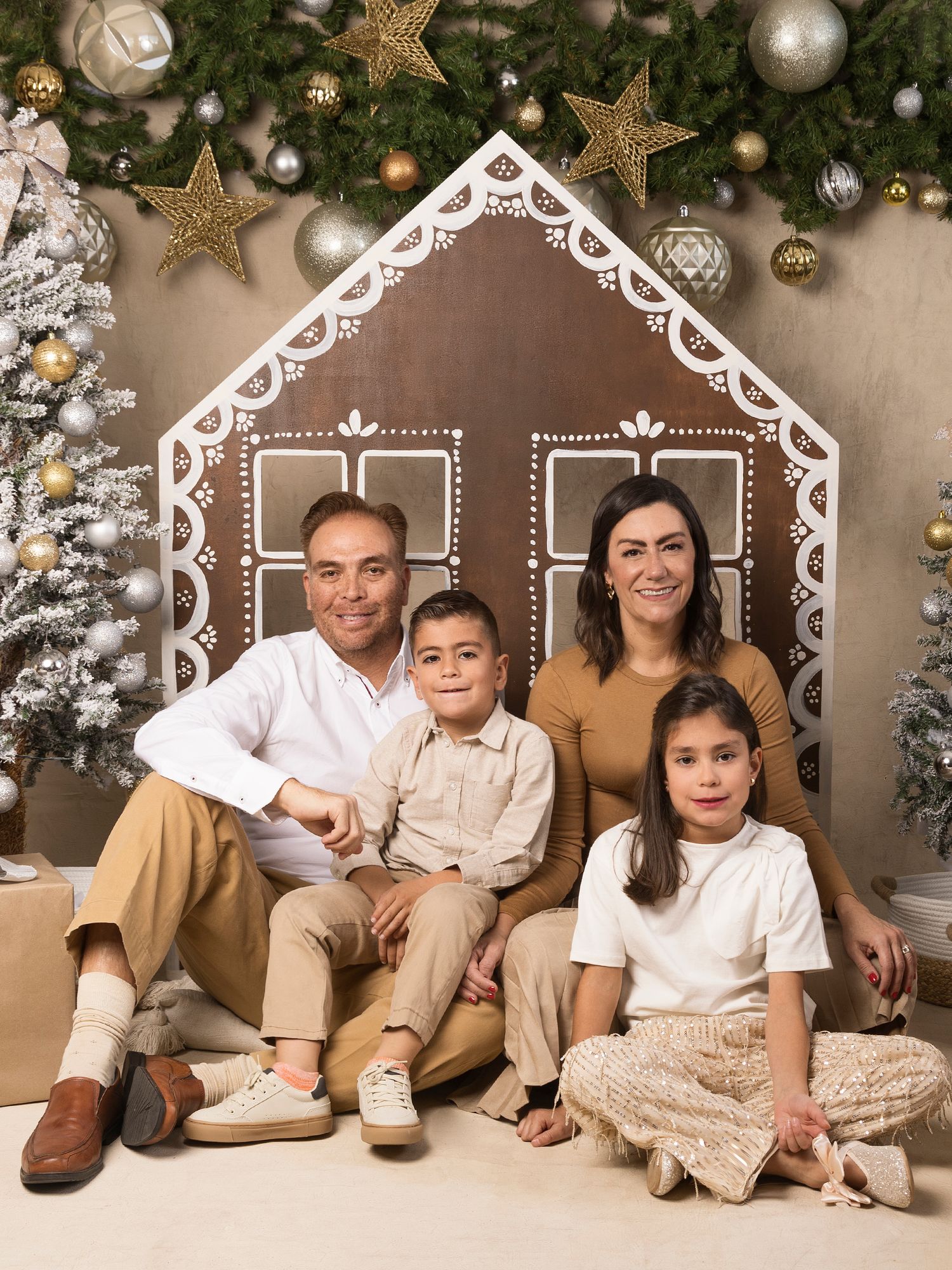 Family Christmas portrait with parents and two children posed in front of a gingerbread house backdrop.