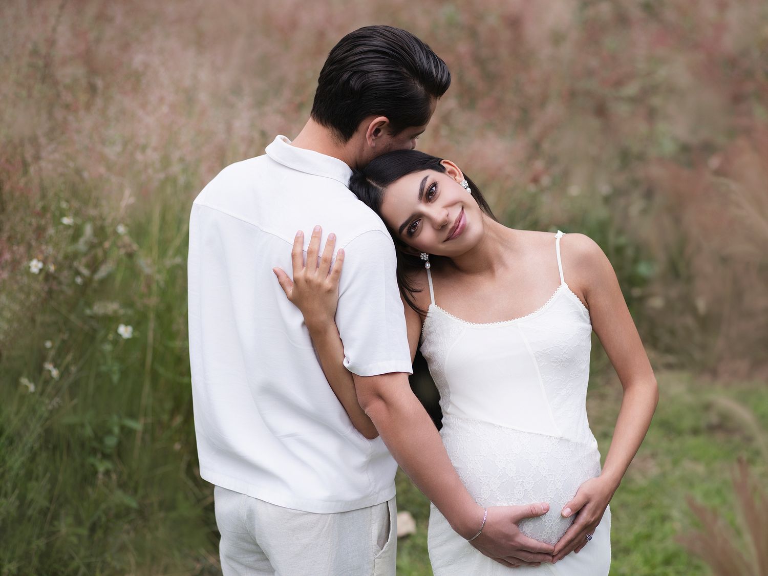 Expecting couple in white outfits embracing tenderly in a field of wildflowers during outdoor maternity session.