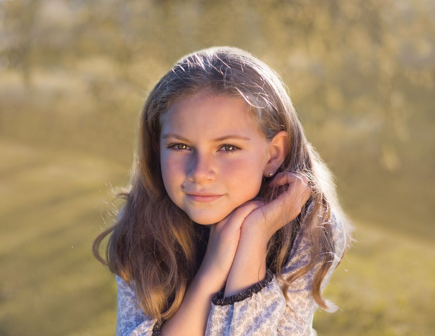 Young girl with long hair posing thoughtfully hands near face in soft golden outdoor portrait.