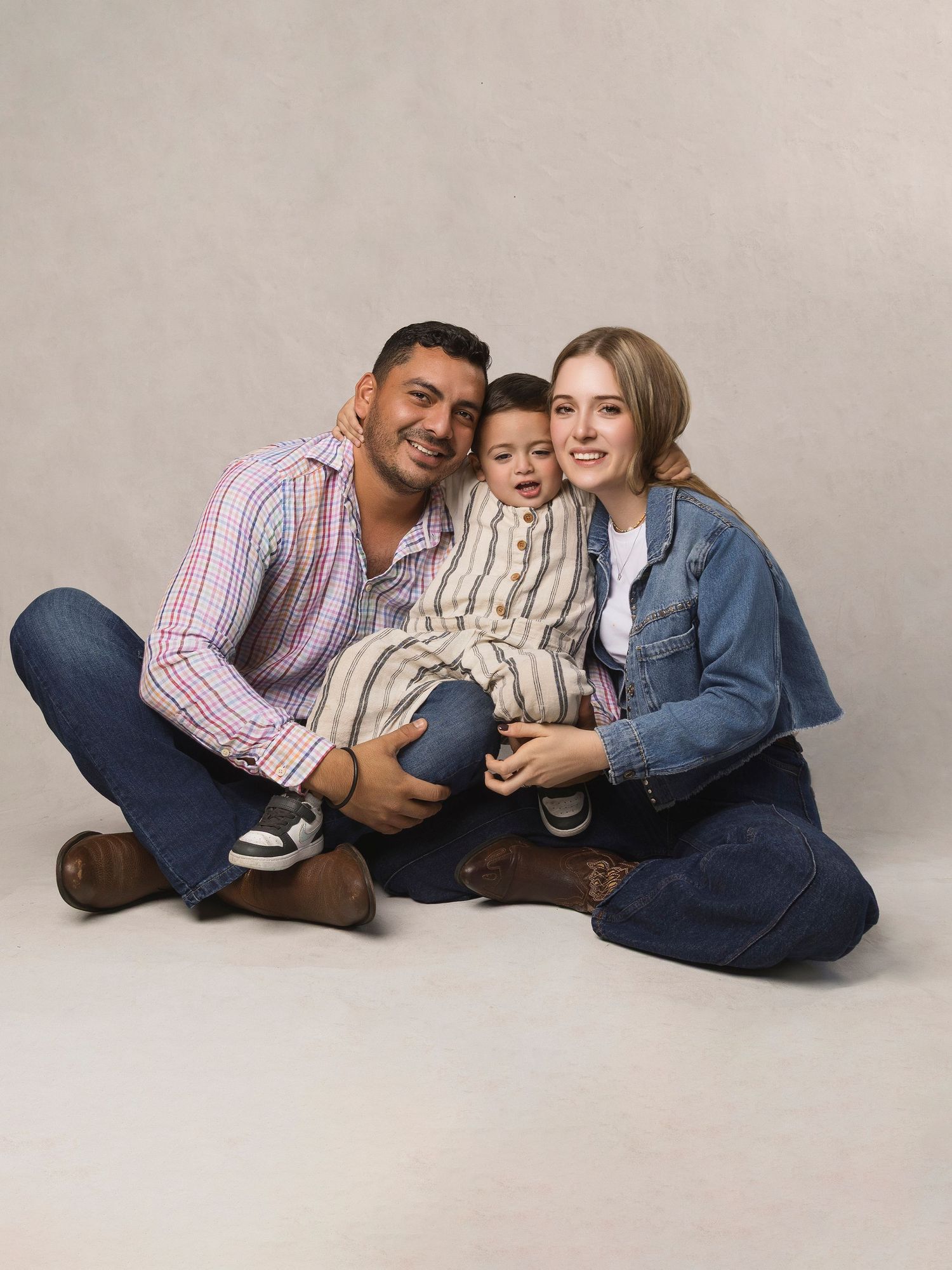 Young family portrait with parents and toddler sitting on floor, smiling warmly in casual denim outfits against neutral background.