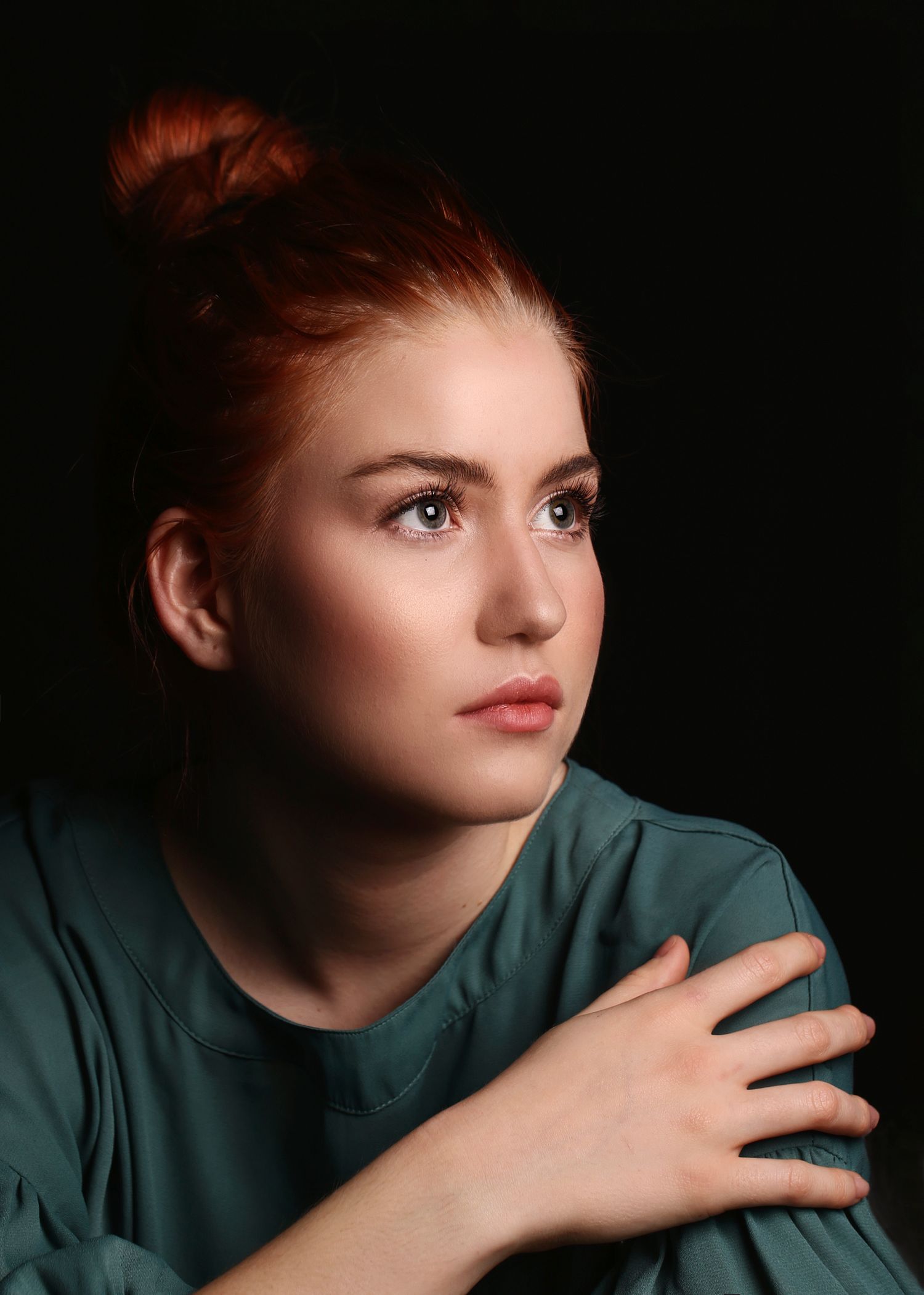 Color portrait of a young redheaded woman in a teal top with hair in a bun on dark background.