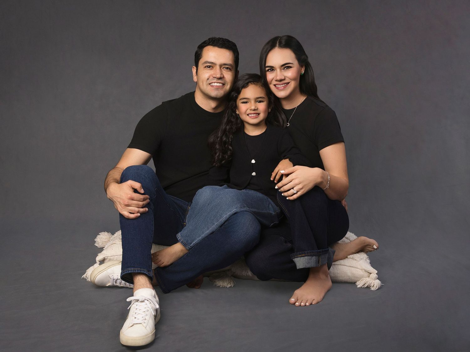 Family of three in matching black outfits sitting casually on studio floor together.
