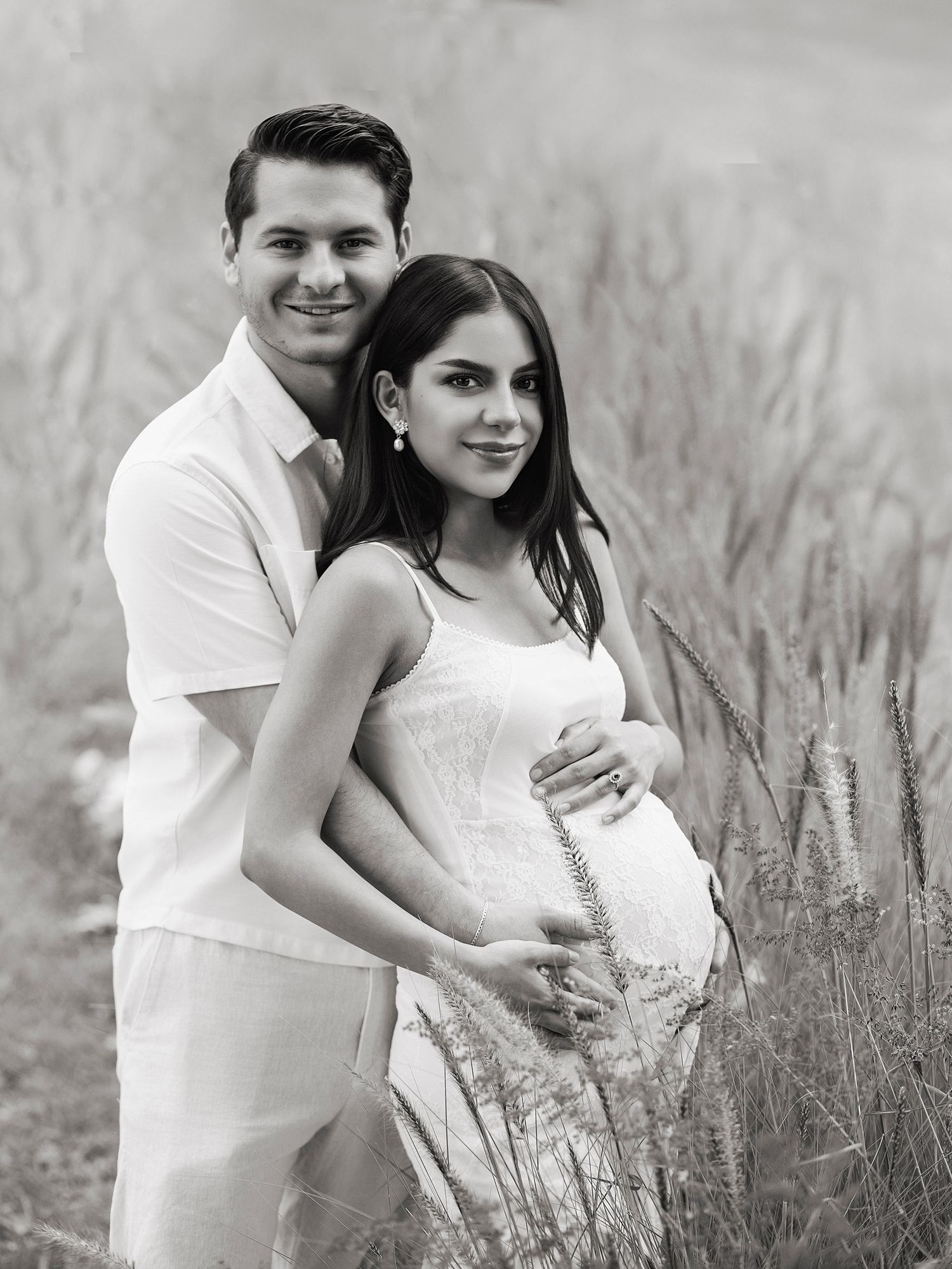 Black and white maternity photo of couple embracing in a field, woman in white dress holding baby bump.