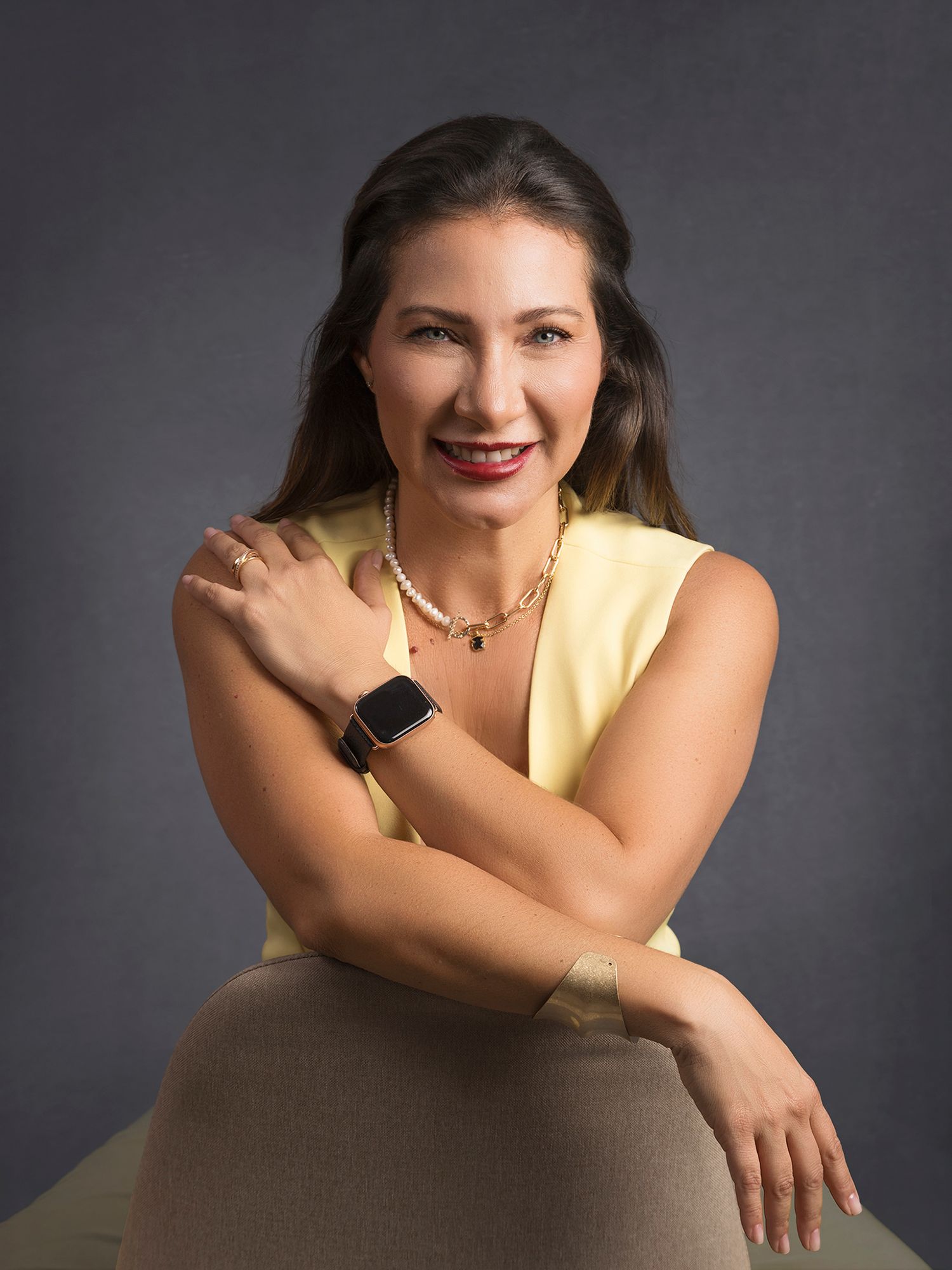 Smiling woman in yellow top with pearl necklace and smartwatch poses in studio portrait.