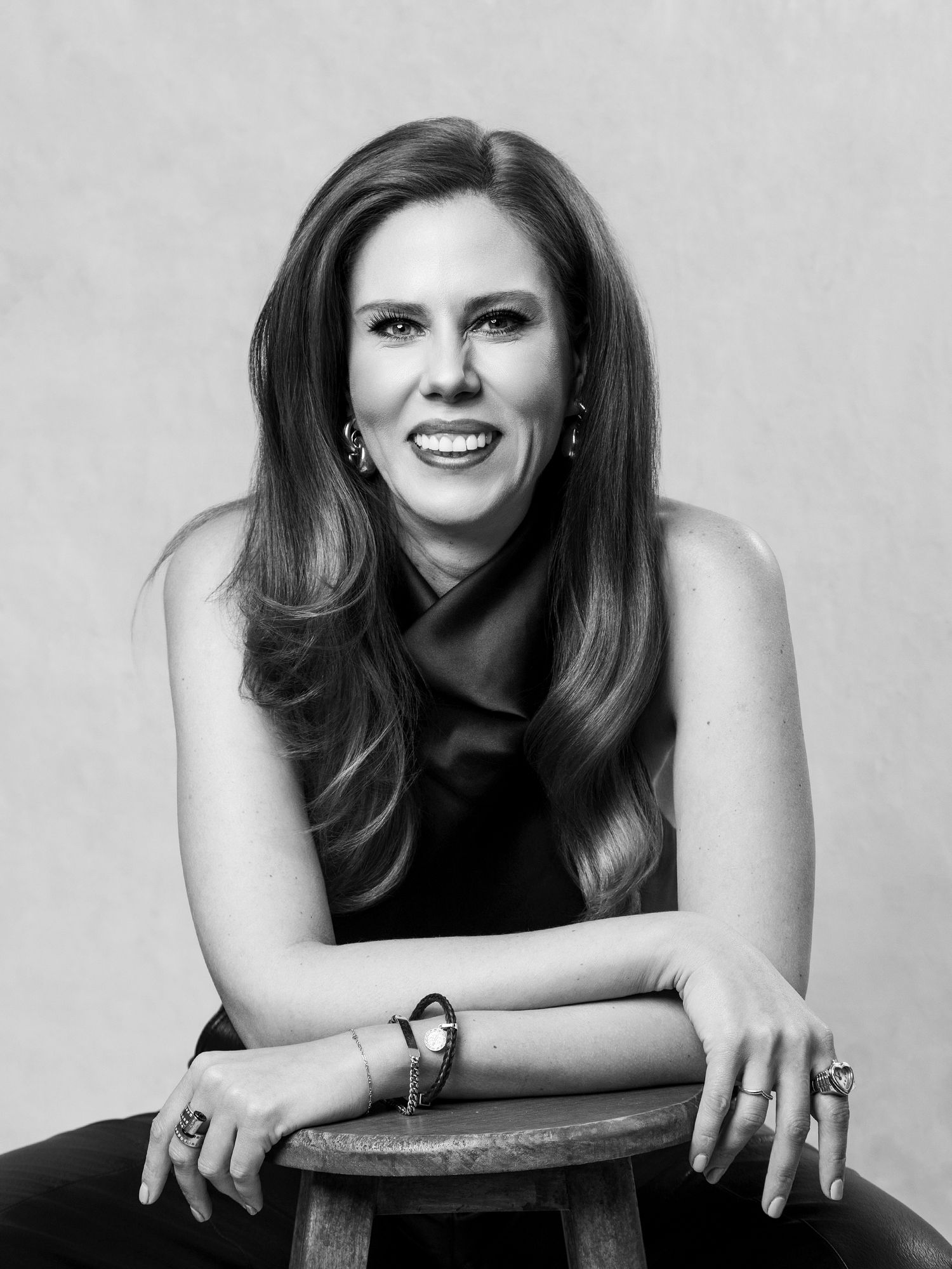 Black and white portrait of smiling woman in sleeveless dress leaning on wooden stool.
