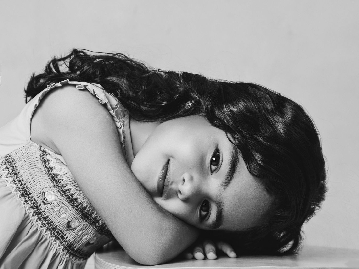 Black and white portrait of young dark-haired girl resting head on surface, smiling softly.