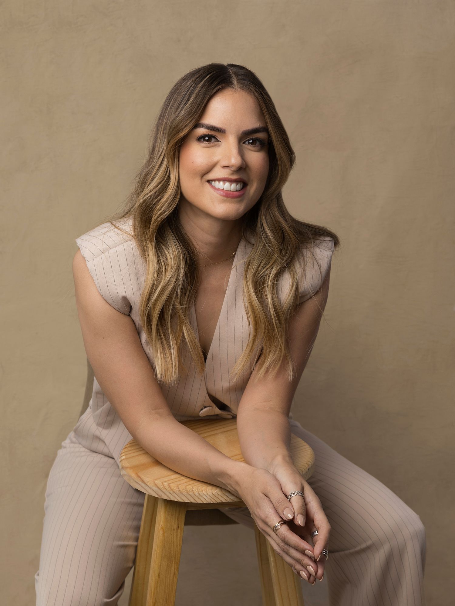 Smiling woman in beige ribbed outfit sits on wooden stool in warm studio portrait.