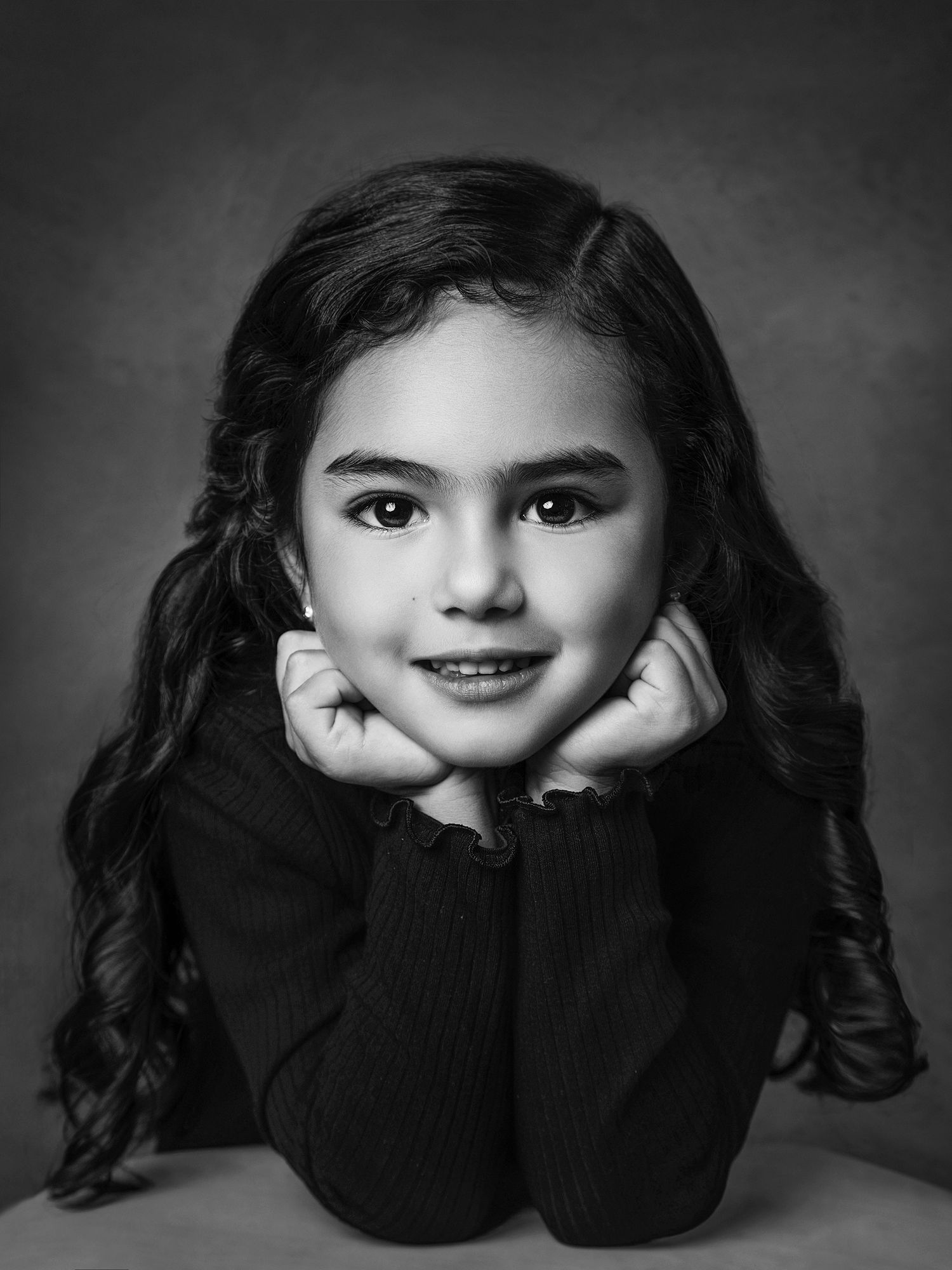 Black and white portrait of young girl with dark hair, resting chin on hands, studio photography.