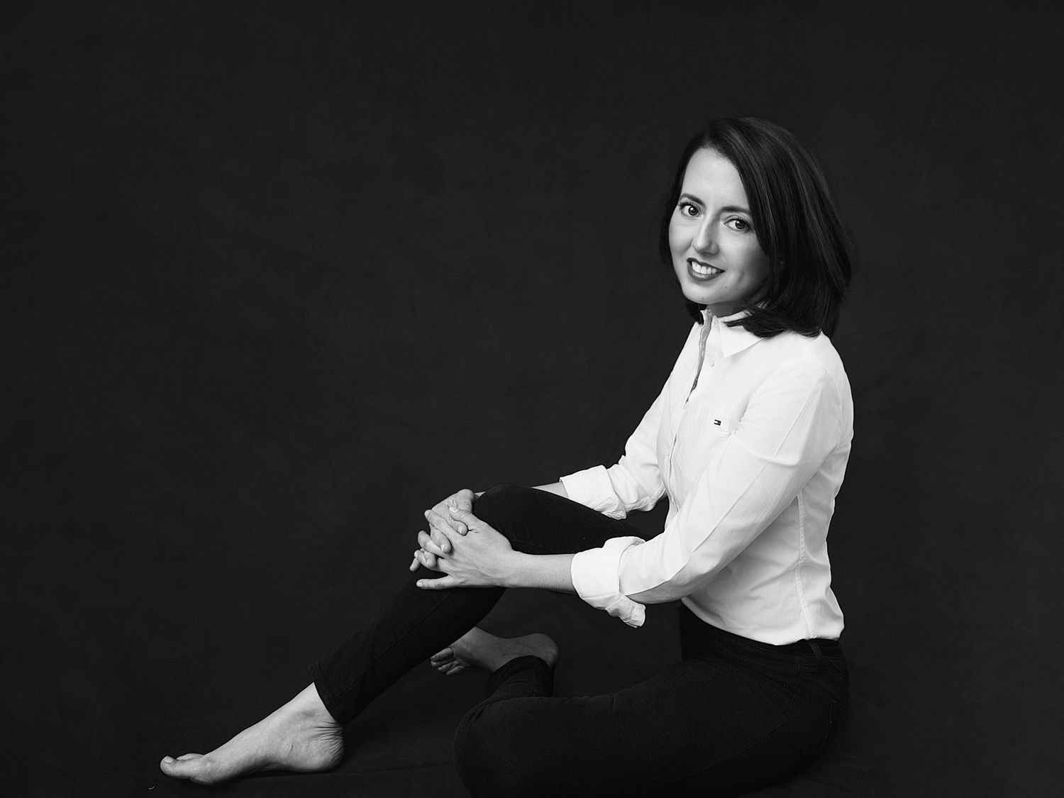 Black and white portrait of barefoot woman in white shirt and dark pants sitting casually on floor.