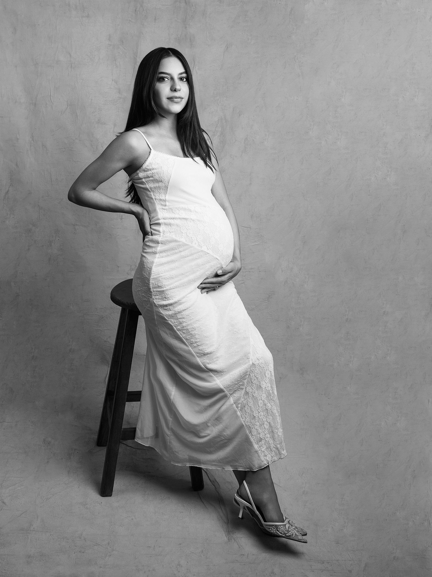 Black and white studio maternity portrait of pregnant woman in white dress leaning against a wooden stool.