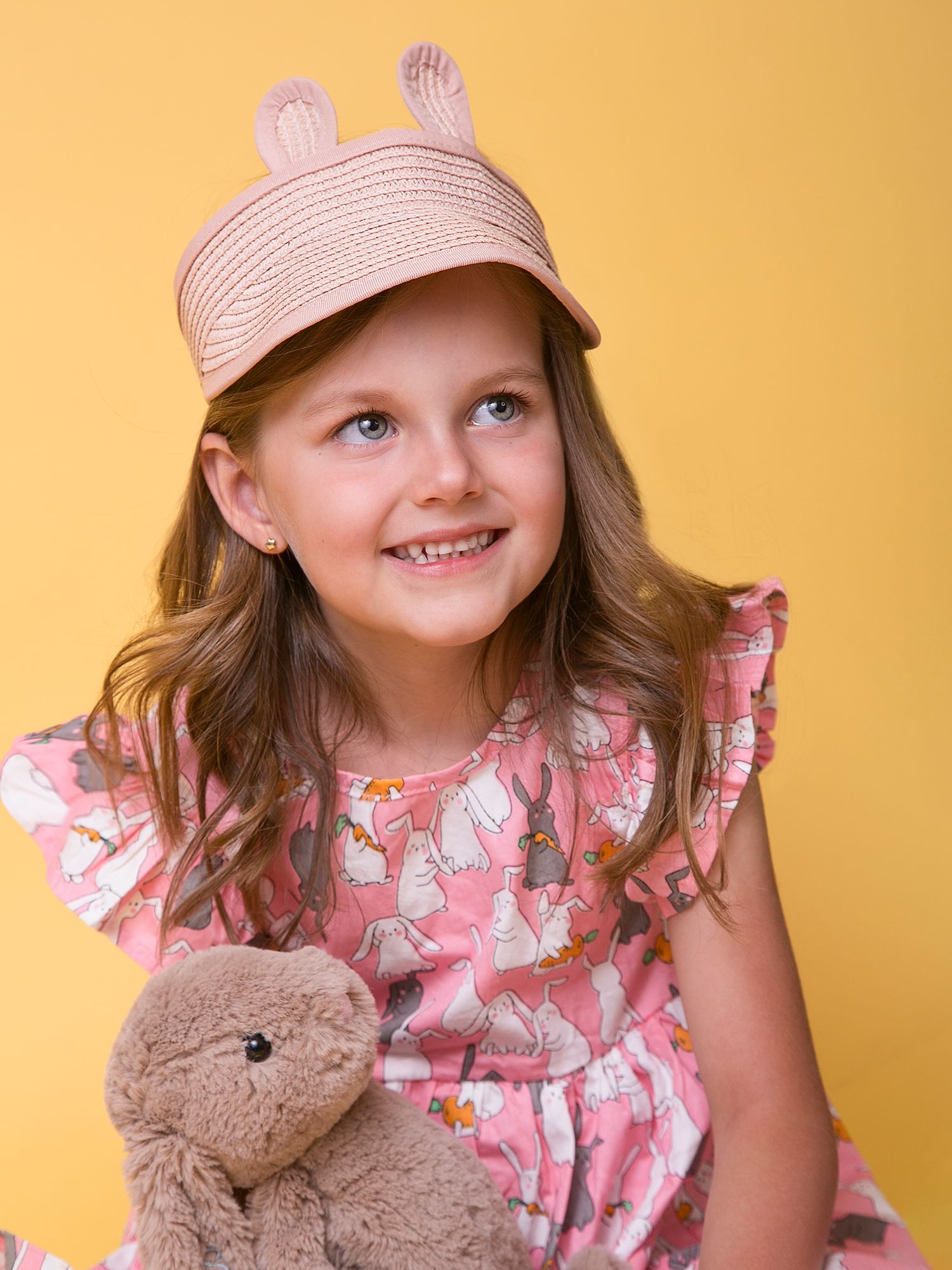 Cheerful girl in pink floral dress wearing bunny ear hat holding teddy bear on yellow background.