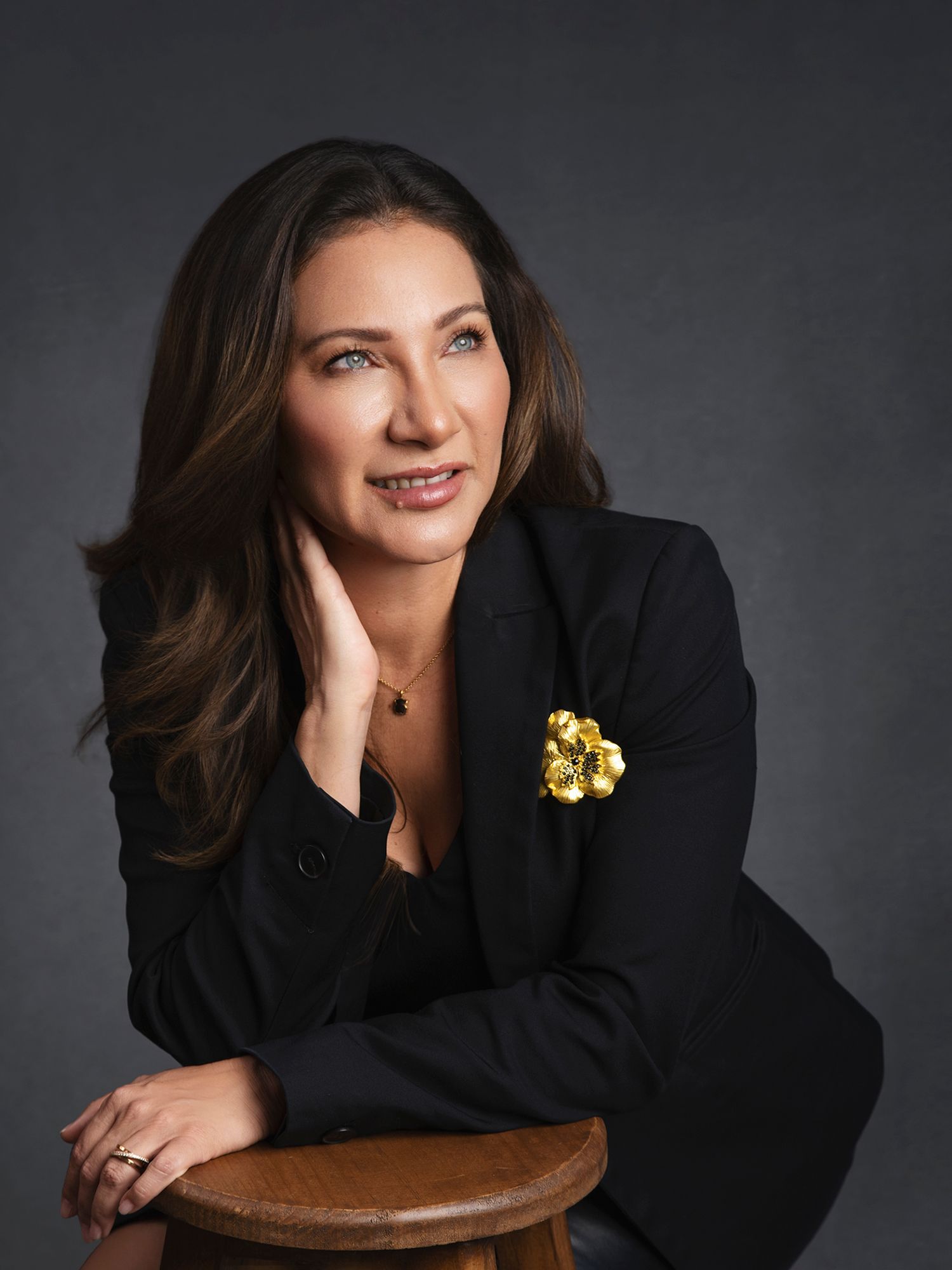 Woman in black blazer with gold floral brooch leans on stool in elegant studio portrait.
