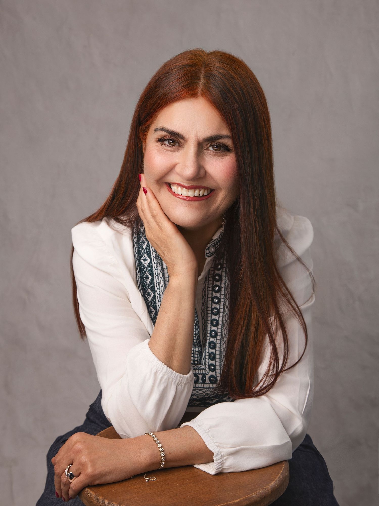 Smiling woman with long red hair poses in white blouse and embroidered vest against gray studio background.