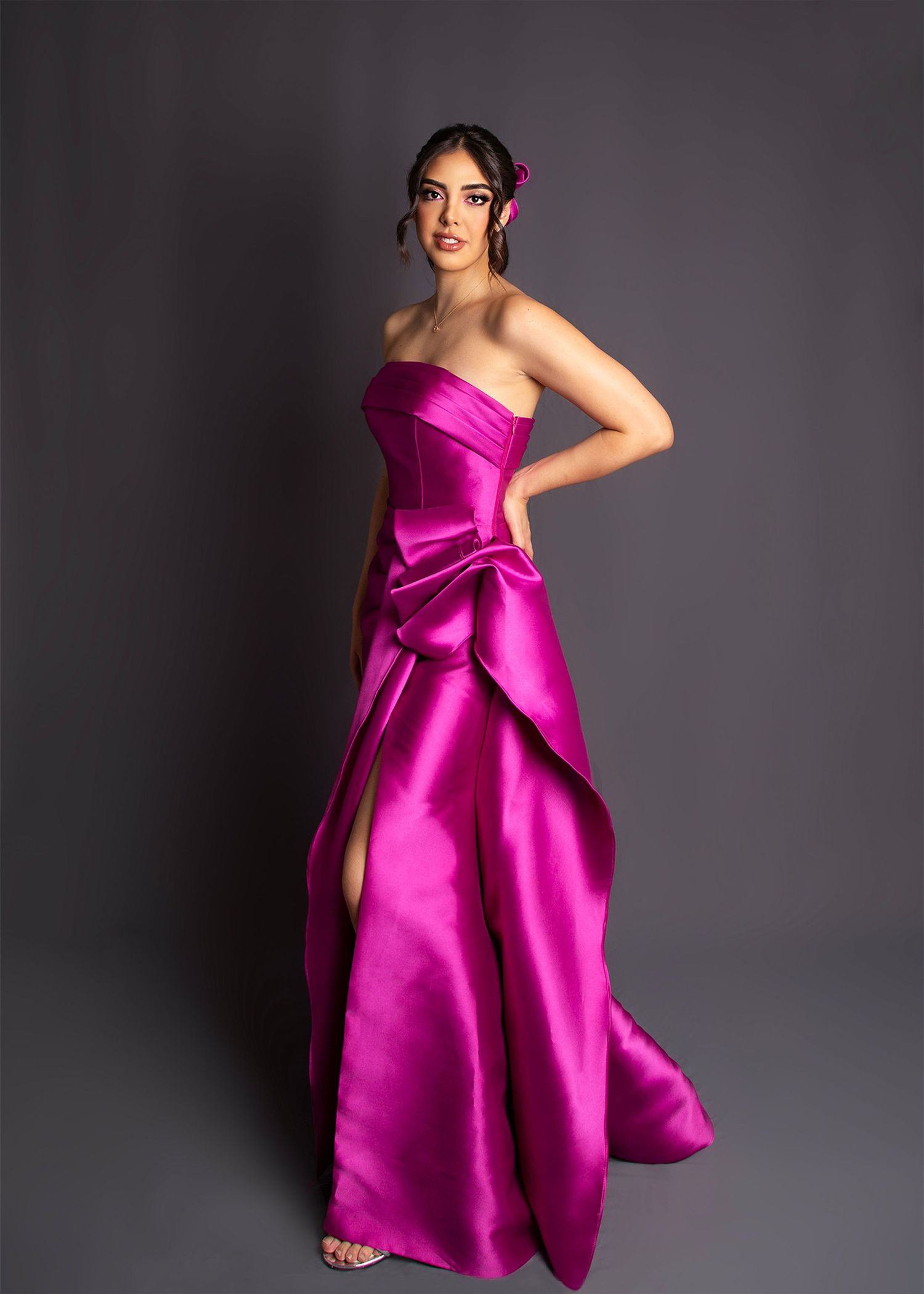 Woman in strapless fuchsia satin ball gown with dramatic bow and high slit posing in studio.